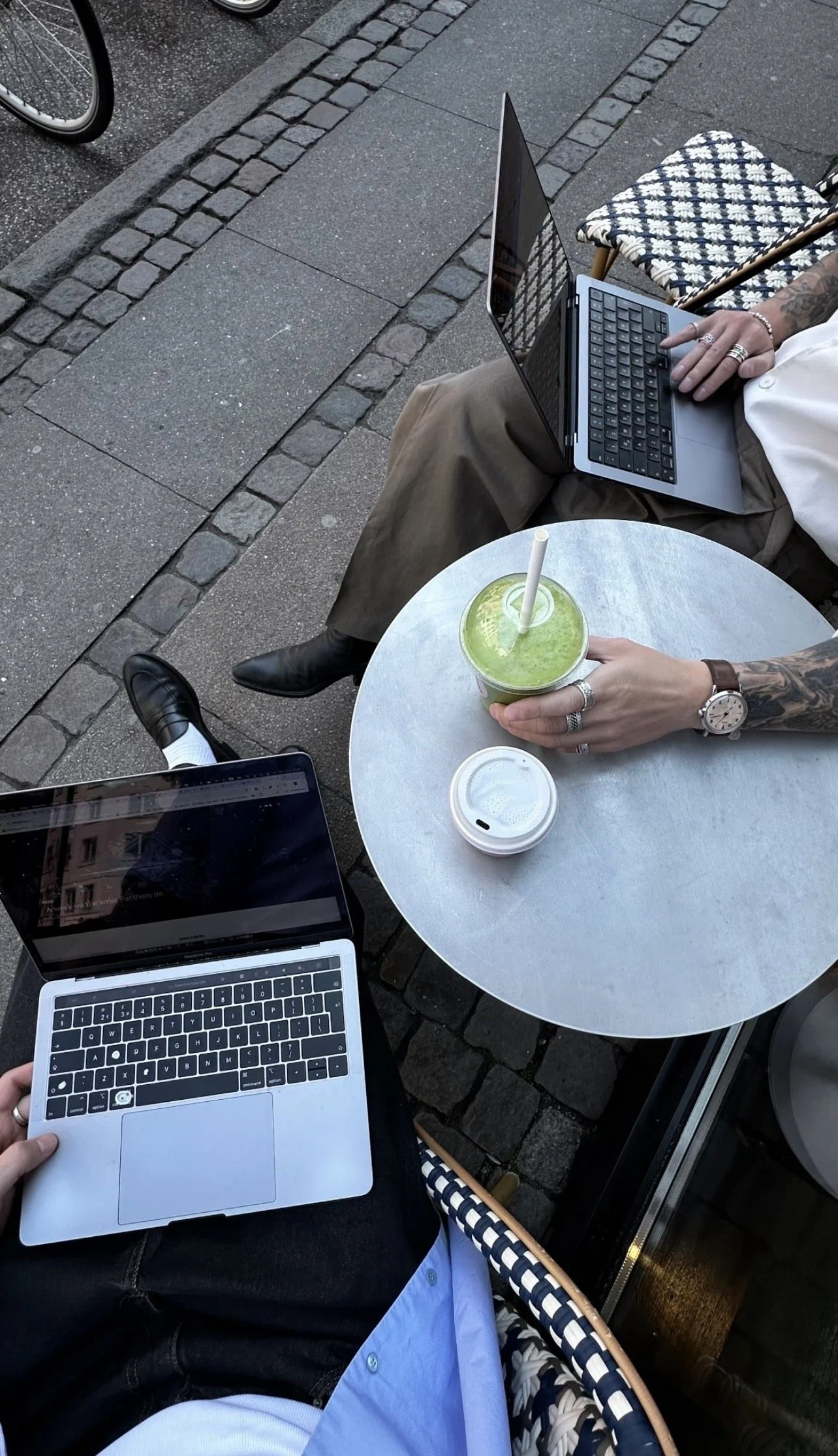 Two people working on laptops at a cafe table outside. One person's hand is visible holding a MacBook, while the other person's hand is on a second laptop. A green smoothie and a coffee cup are on the table.