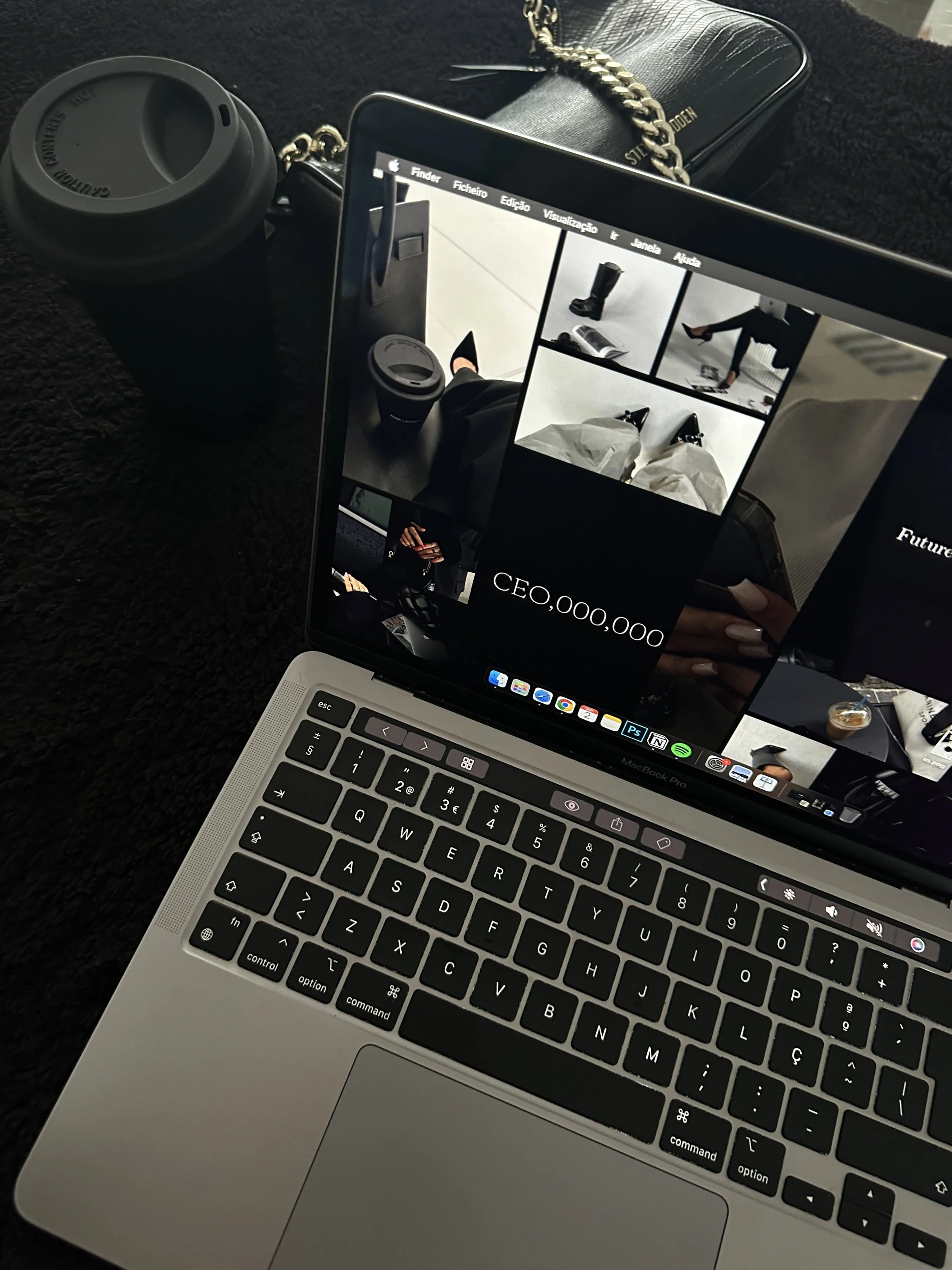 A laptop on a dark surface displaying a collage of fashion-related images and a partially visible cup of coffee or tea, surrounded by a black purse with a chain strap and a black travel mug with a lid.