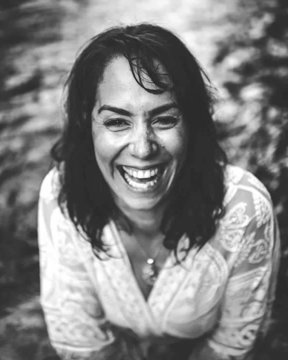 A woman with dark, curly hair laughing with a wide smile, outdoors.