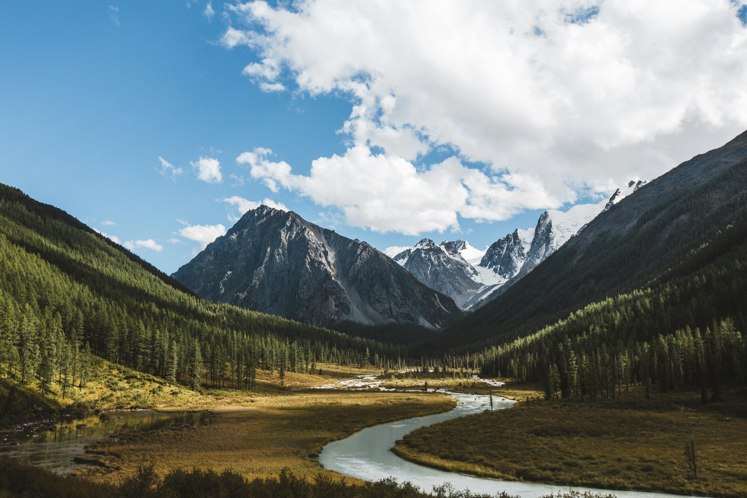 A scenic landscape of a river flowing through a flat valley surrounded by green pine forests and tall snow-capped mountains under a partly cloudy blue sky.