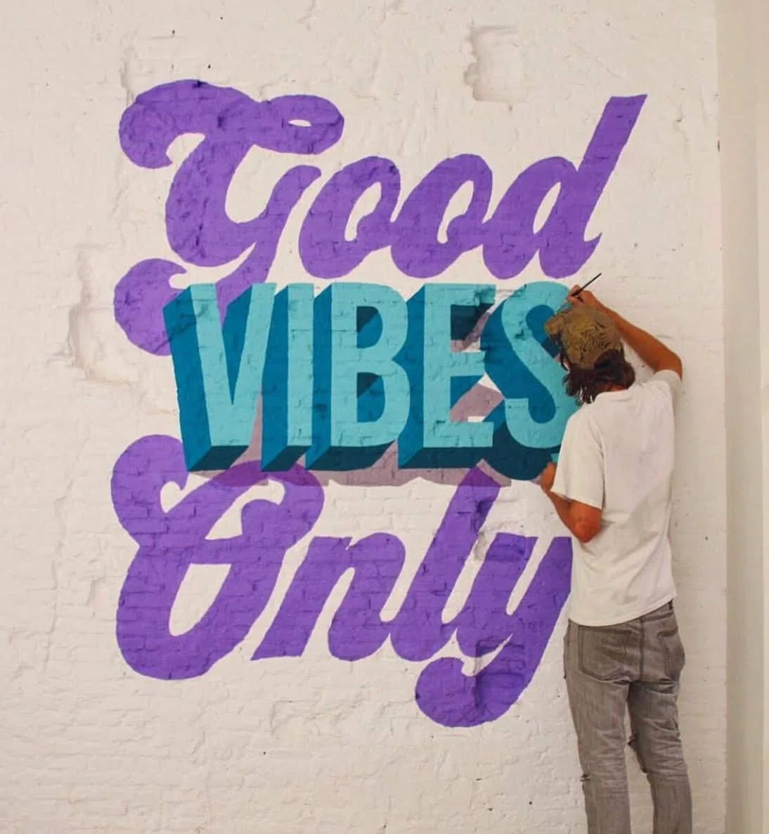 Artist painting 'Good Vibes Only' mural on white brick wall
