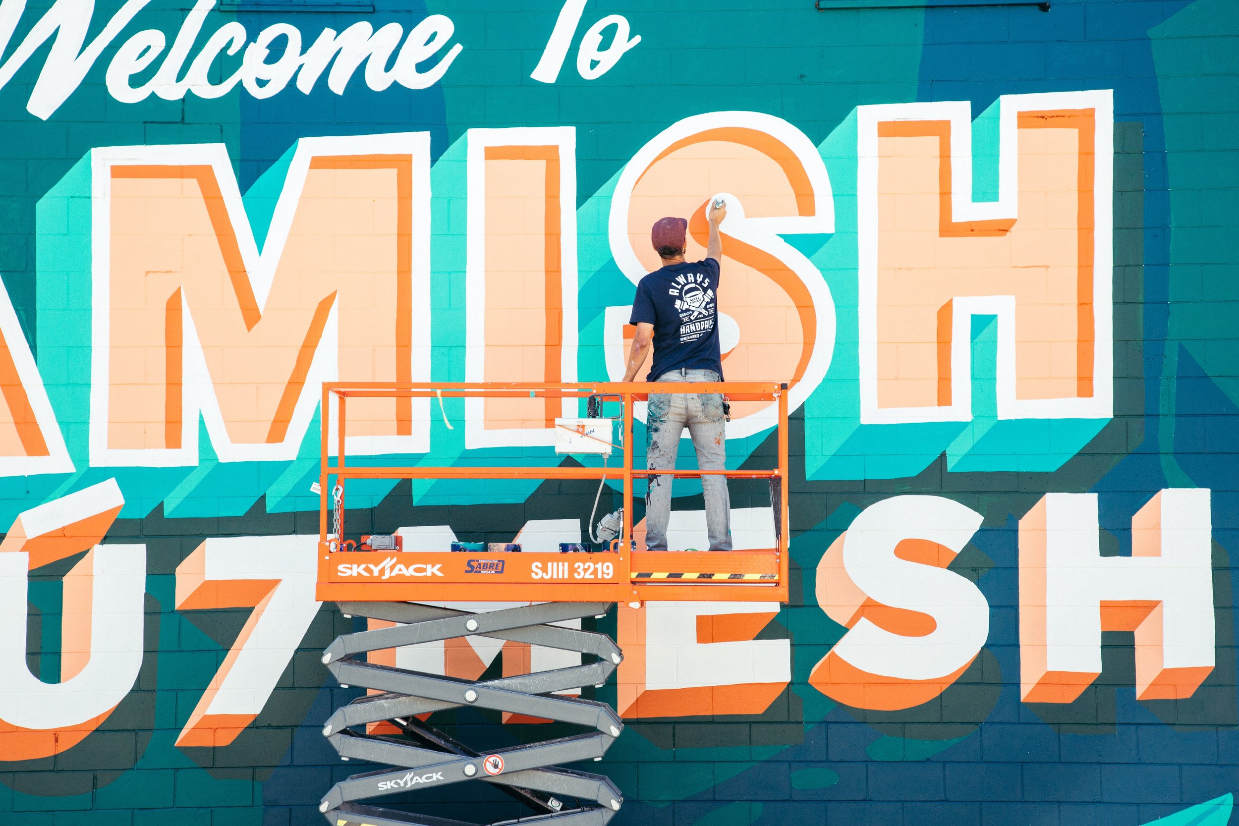 Street artist painting a large colorful mural on a wall using a scissor lift.