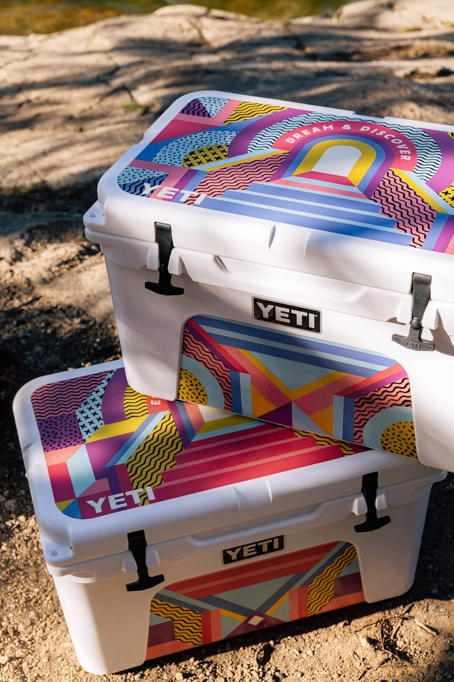 Two YETI coolers featuring colorful geometric patterns, set outdoors on a sandy surface.