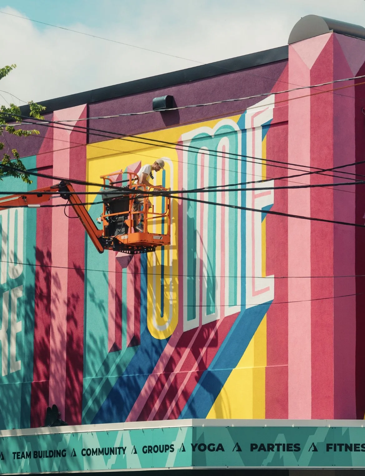 A person on a cherry picker painting a colorful mural on the side of a building with bold letters. The building's awning features words such as 'TEAM BUILDING' and 'YOGA.' Overhead utility lines and tree branch shadows are visible.