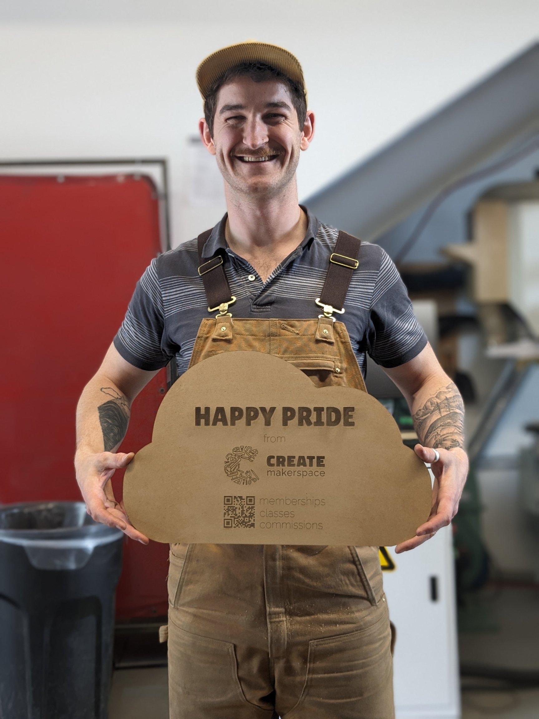 Person holding a wooden cloud-shaped sign that reads 'Happy Pride from Create Makerspace' with QR codes for memberships, classes, and commissions, standing in a workshop environment.