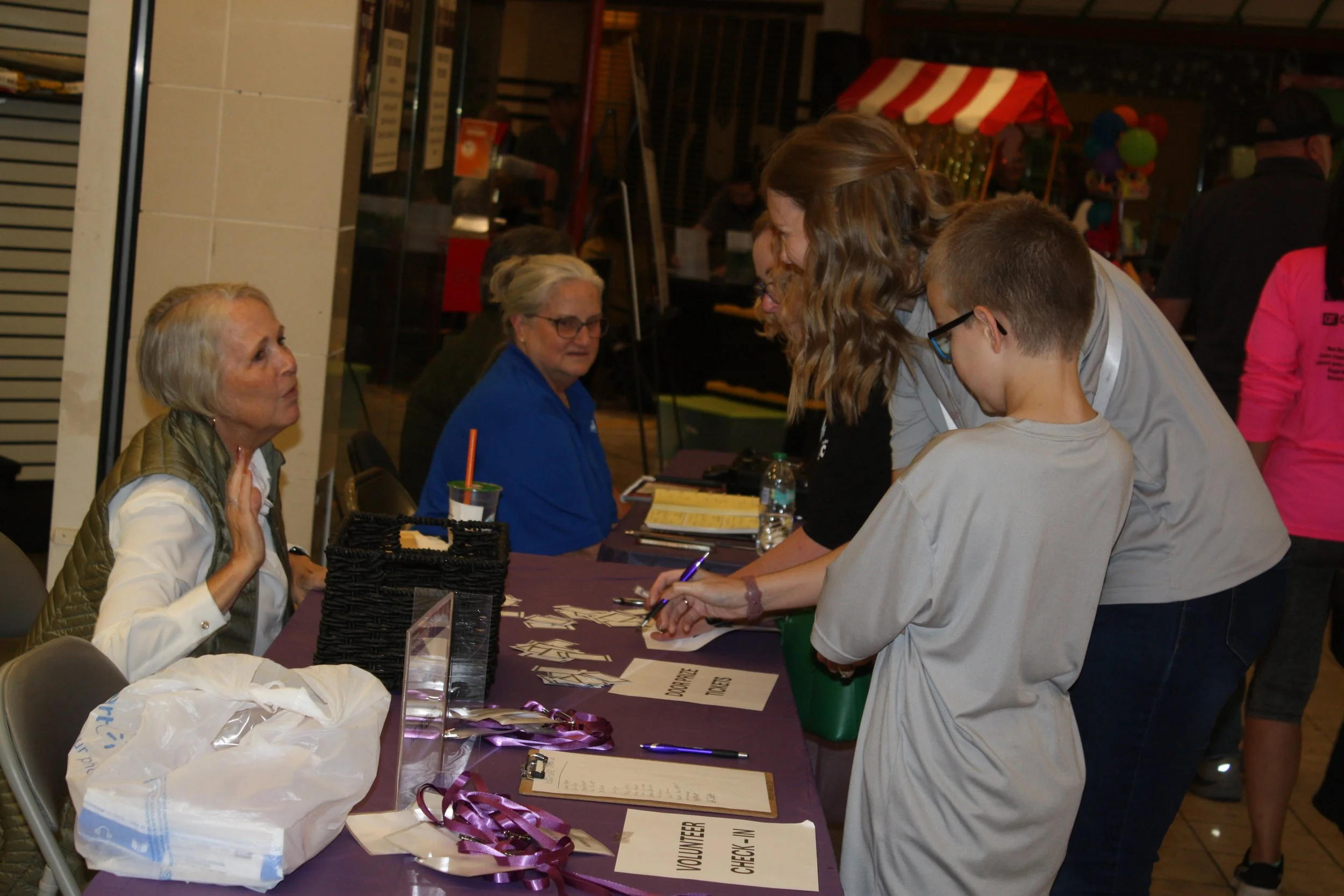 People at a volunteer sign-in table, including older women and younger individuals, with signs indicating volunteer check-in, in an indoor setting with balloons and possibly a fair or community event background.