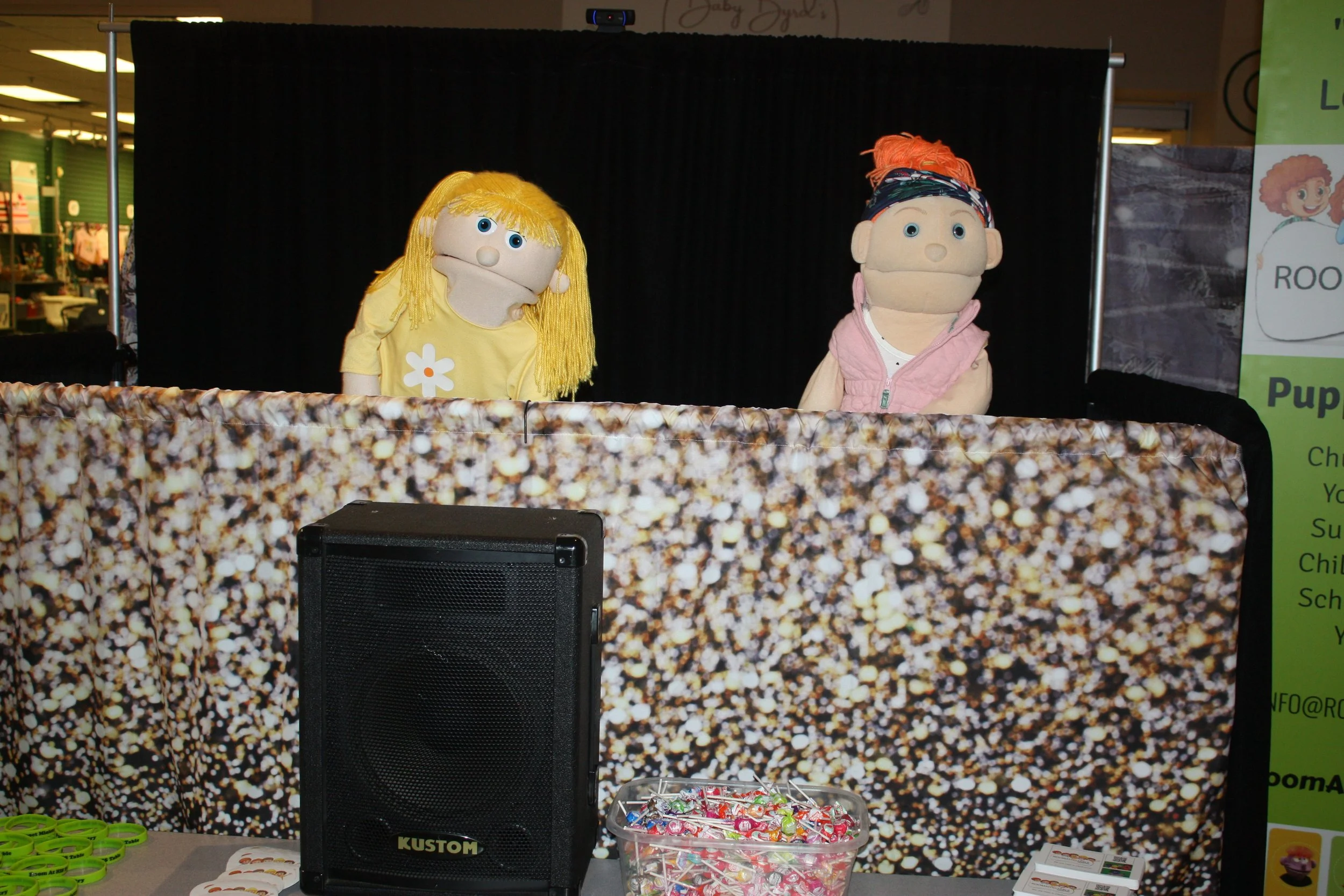 Two puppets, a girl with yellow hair and a boy with orange hair, performed on a stage with a black background at a public event, with candy and informational materials on a table in front.