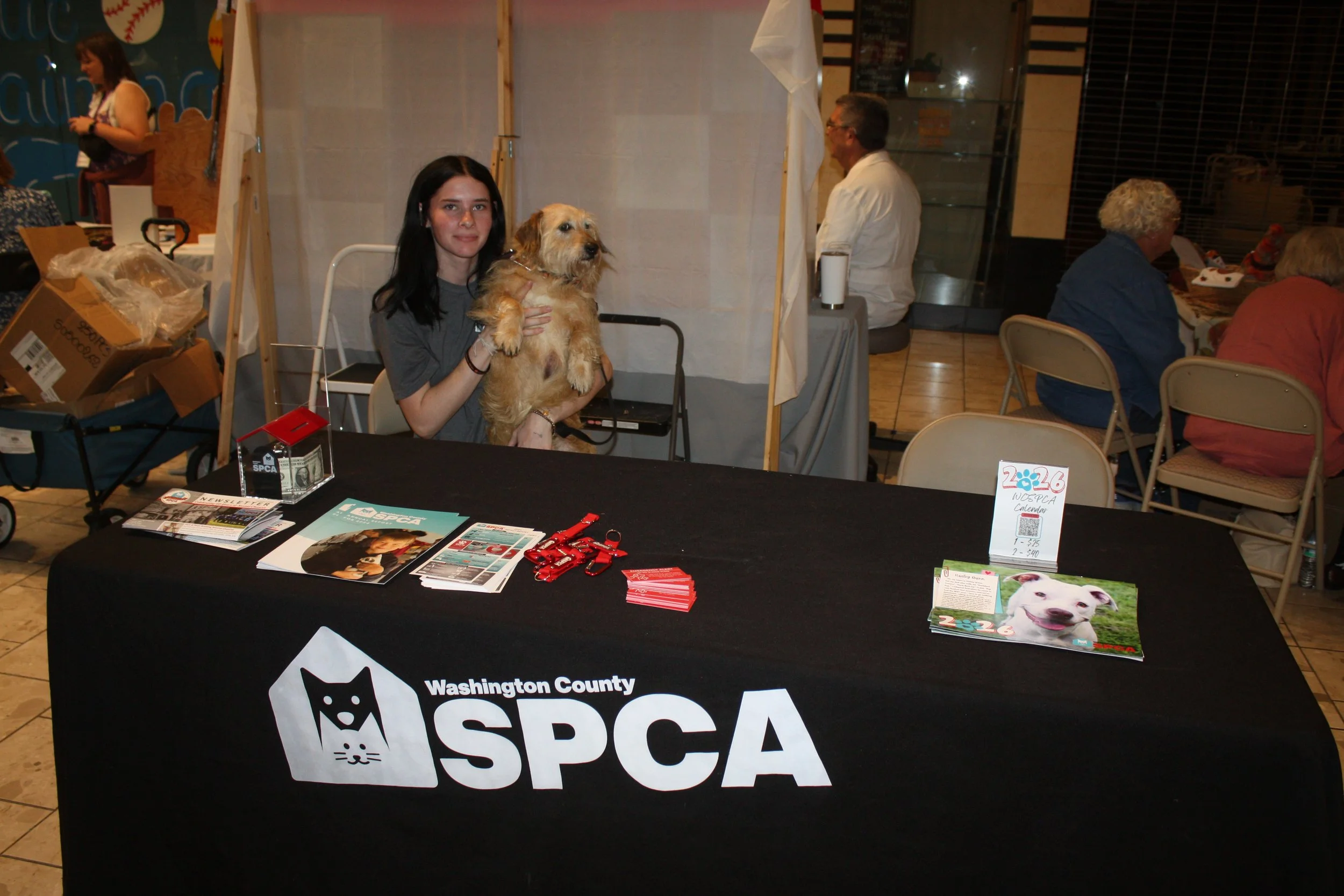 A woman holding a dog at an event booth for the Washington County Humane Society. The booth has brochures and red keychains on a table with a Washington County SPCA banner.