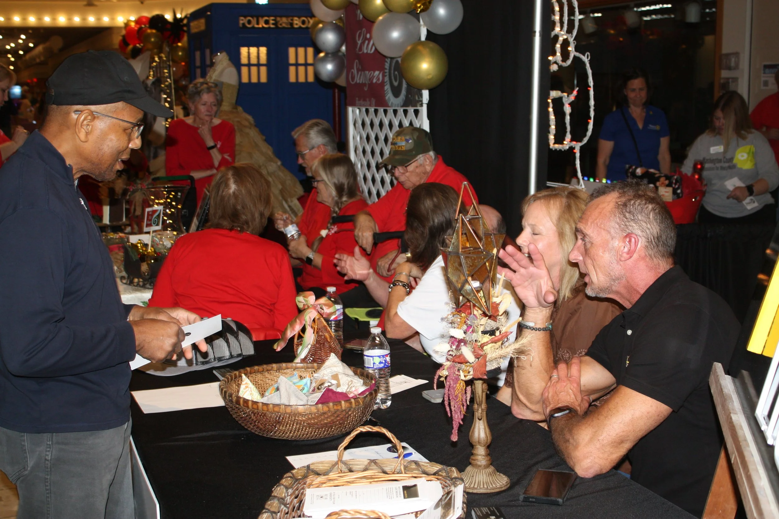 People gathered at a booth at a festive event, possibly a holiday market, with Christmas decorations, balloons, and a table of items for sale or display.