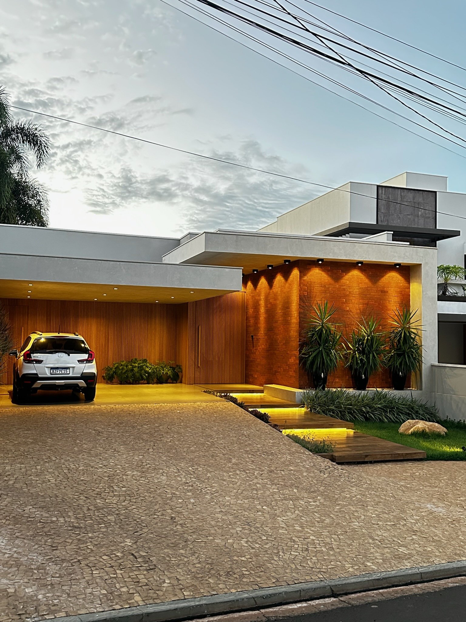 Casa moderna com fachada iluminada à noite, carro estacionado na garagem e jardim com plantas altas.