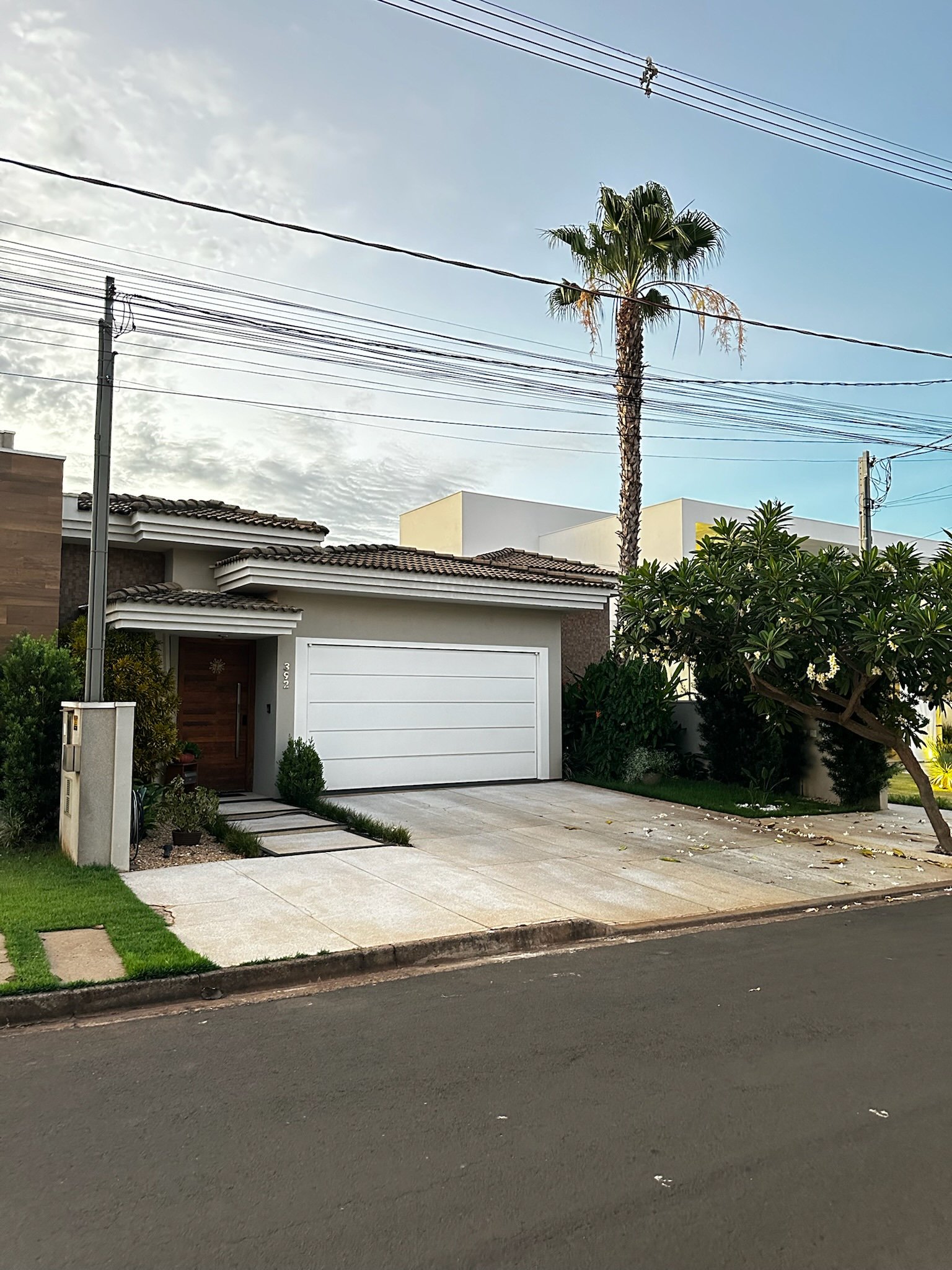 Casa moderna com garagem branca e telhado de telhas, árvore ao lado e postes com fiação elétrica.