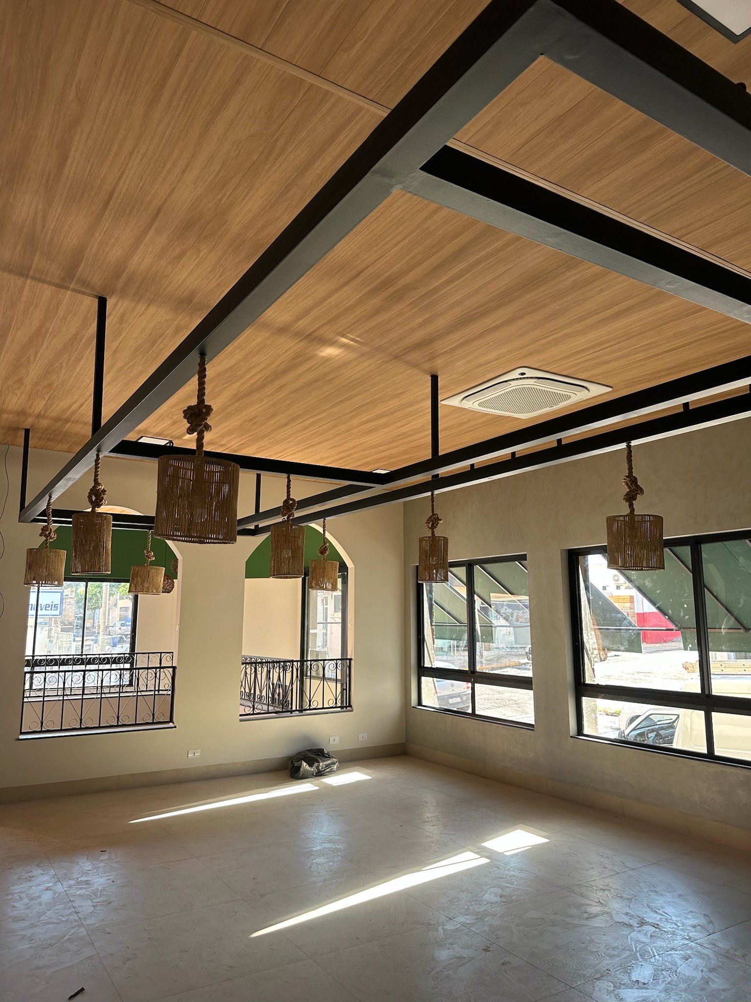 Sala vazia com janelas grandes, teto de madeira com luminárias pendentes e ar-condicionado.