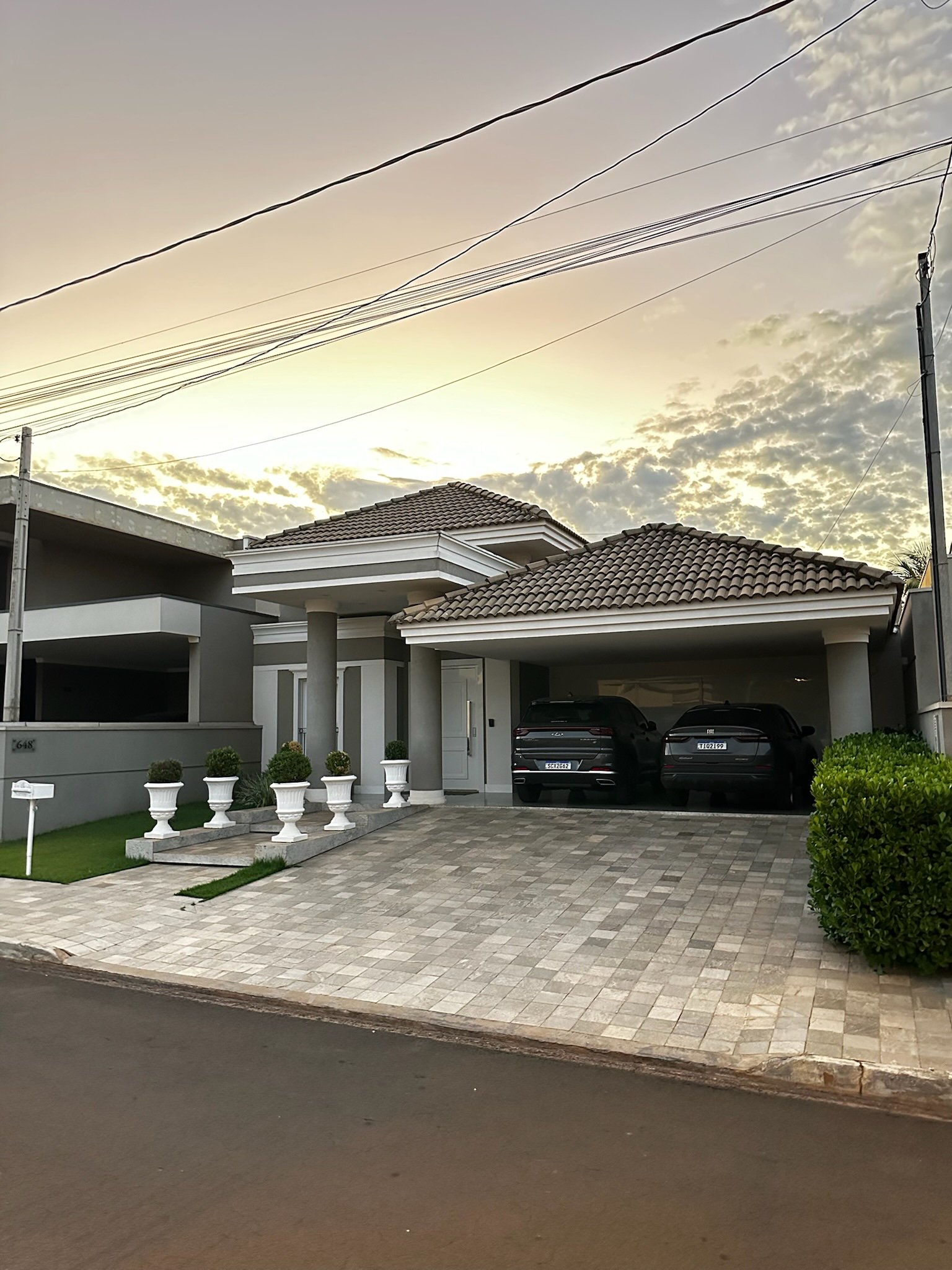 Casa moderna com garagem dupla, dois carros estacionados, telhado de telhas cerâmicas, jardim com vasos decorativos e céu ao entardecer.