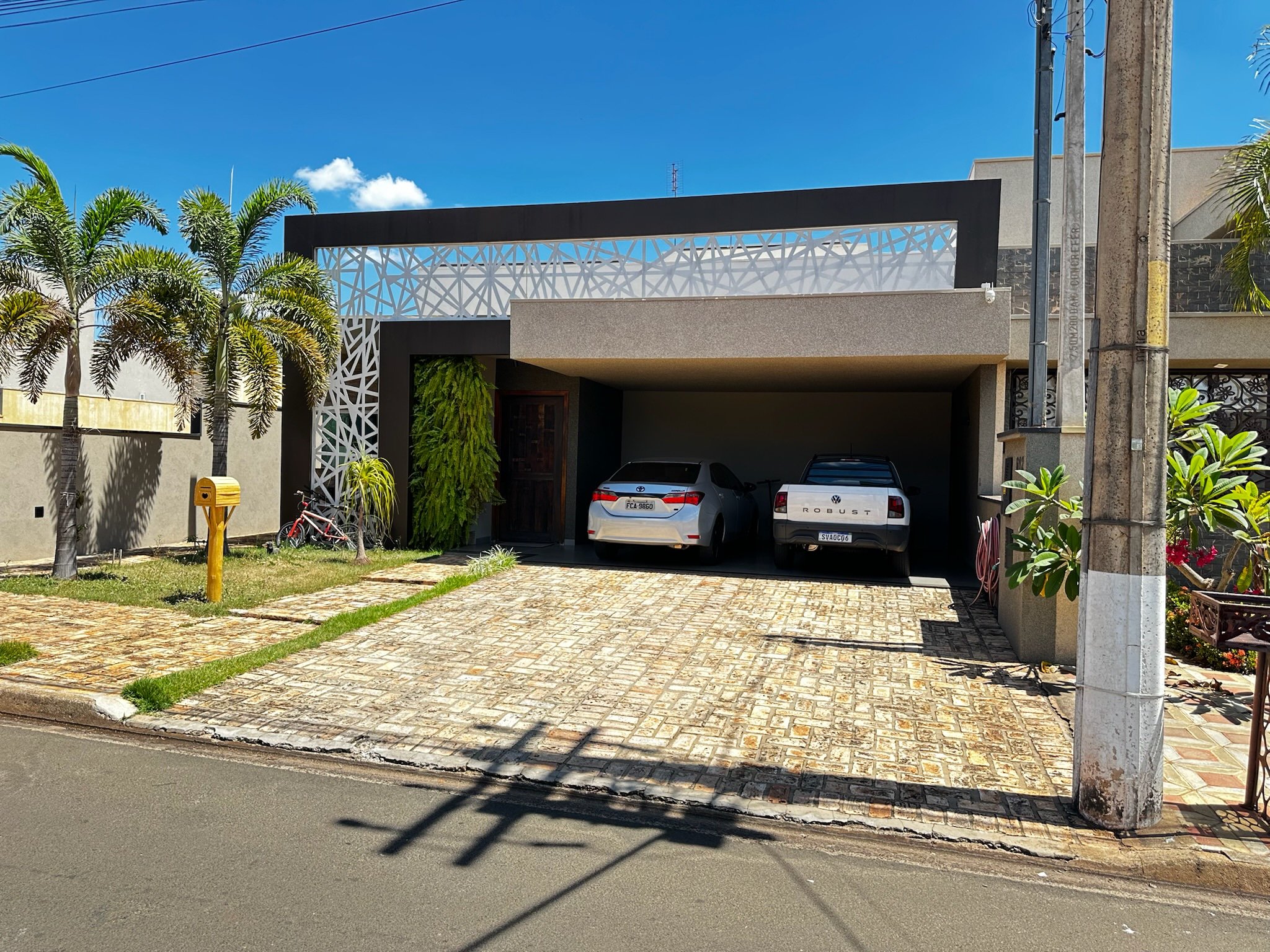 Casa moderna com garagem contendo dois carros, uma picape e um sedan. Jardim com grama, palmeiras e uma bicicleta vermelha ao lado de uma caixa de correio amarela. Estrutura com detalhes geométricos na fachada.