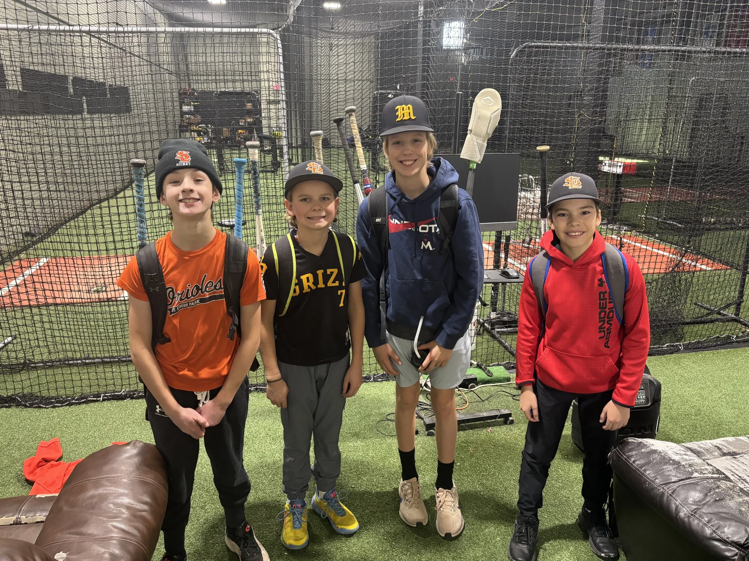 Four boys standing inside a sports facility with batting cages and baseball bats in the background.