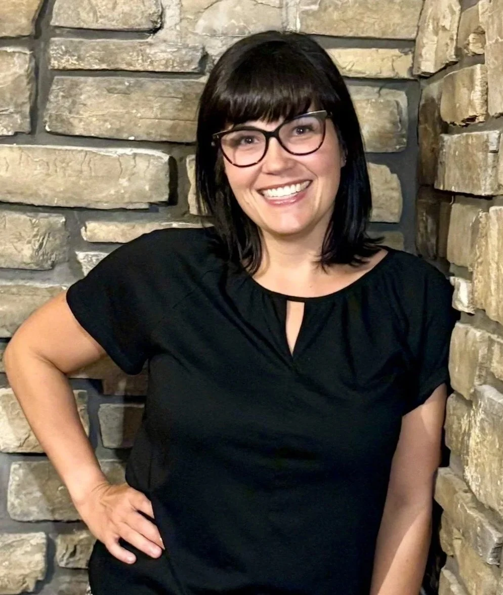 A woman with dark hair and glasses smiling while standing against a stone wall.