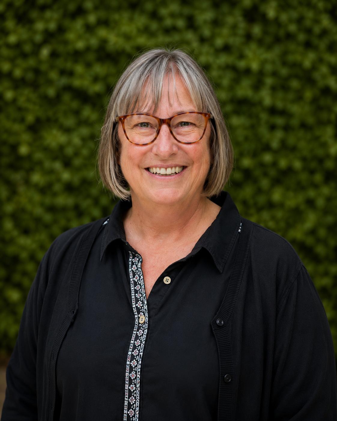 Woman with short, grey hair smiling in front of greenery