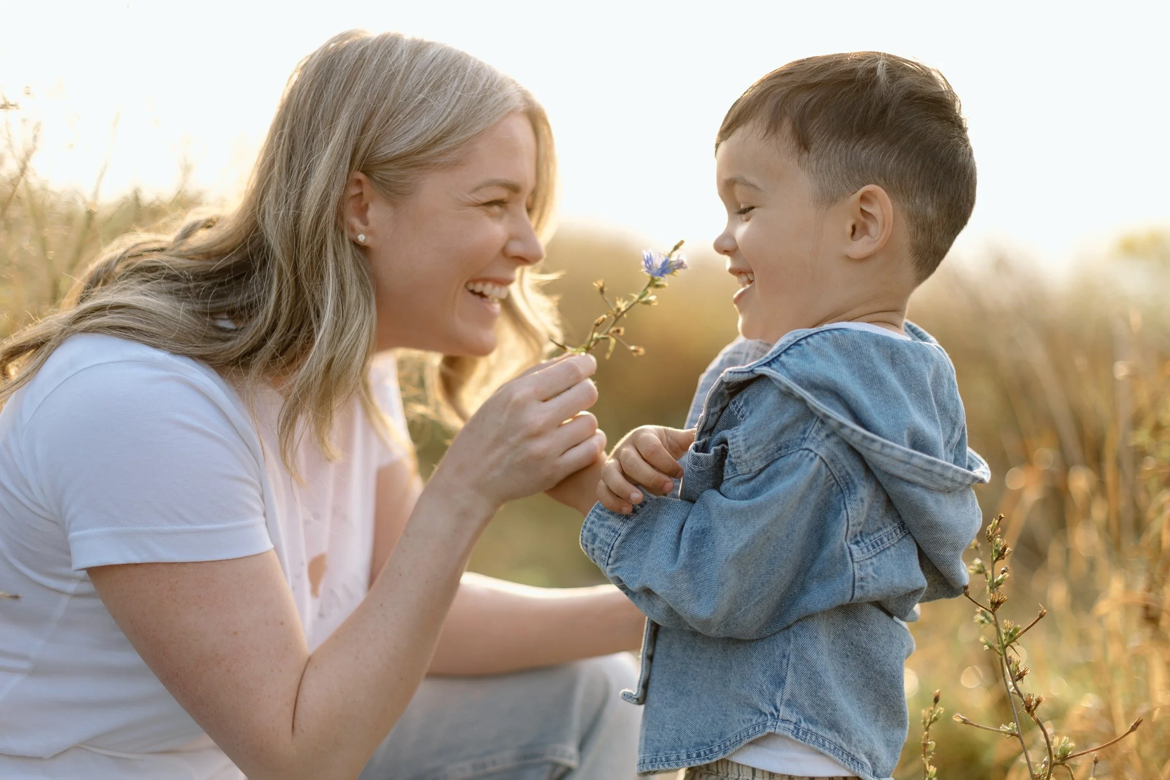 A woman and young boy smiling and playing with a flower in a field during sunset.