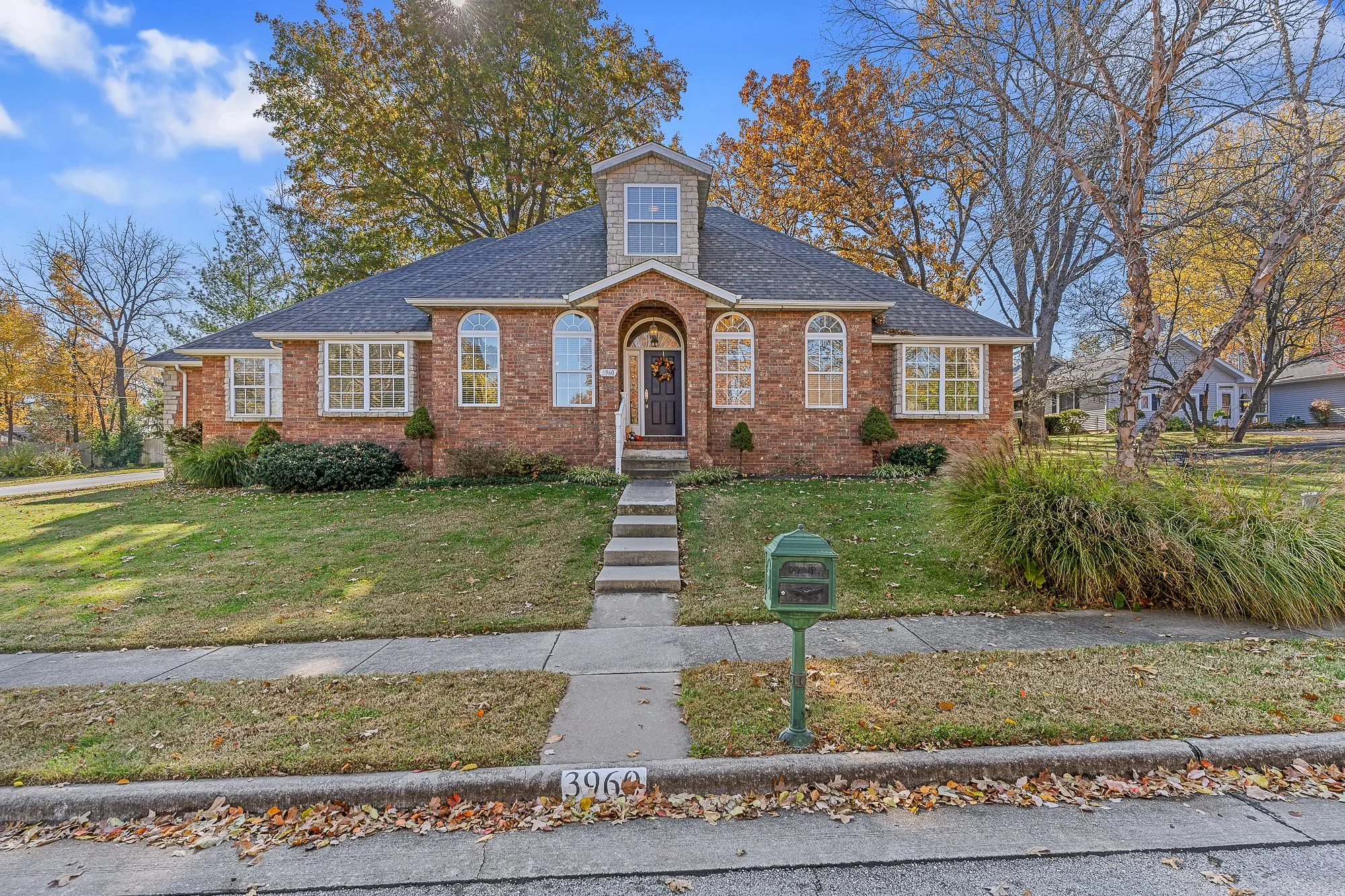 A brick house with a dark front door decorated with a wreath, large arched windows, and a dormer window on the roof. The house is surrounded by trees with autumn foliage and a well-maintained lawn. A mailbox with the number 3961 is in the foreground.