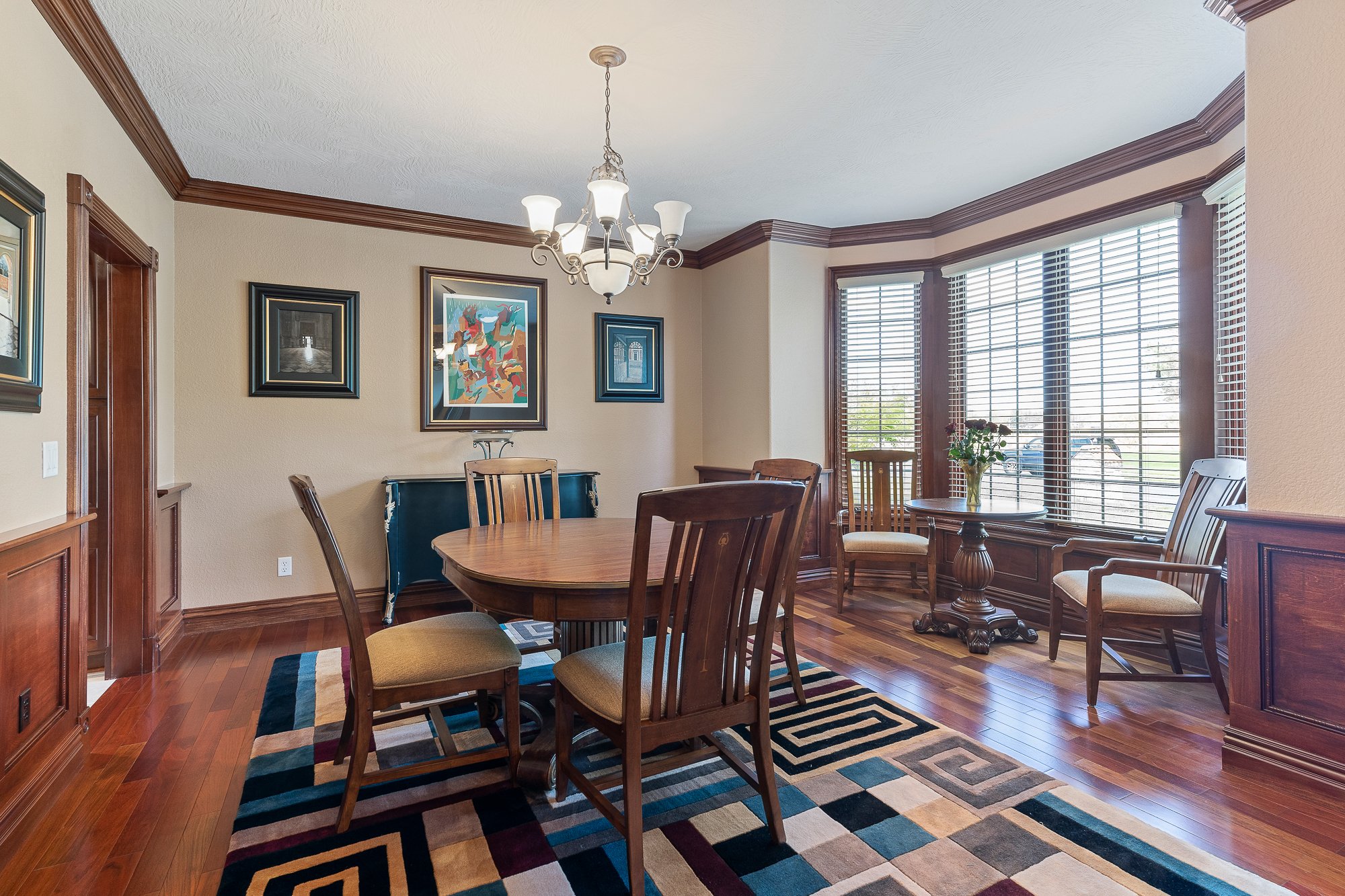 A dining room with a round wooden table, six wooden chairs with beige cushions, a colorful area rug, and a large window with blinds. The room has beige walls, wooden crown molding, and several framed artworks on the wall. A chandelier hangs from the 