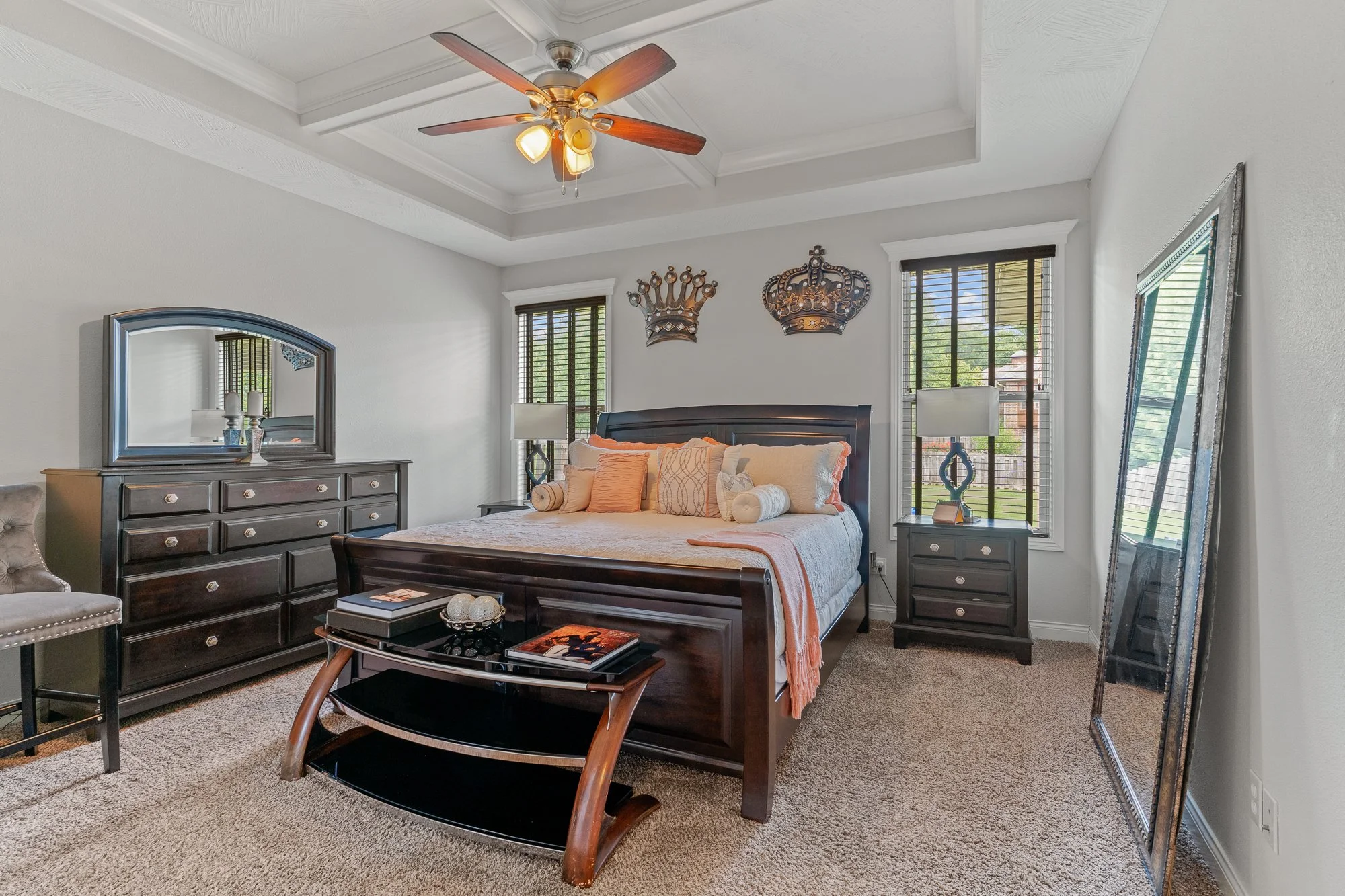 A bedroom with a dark wood bed, nightstands, a dresser with a mirror, and a large mirror leaning against the wall. The bed has beige and orange pillows, a tufted chair, and two lamps. There are windows with blinds, and decorative crown and crown wall