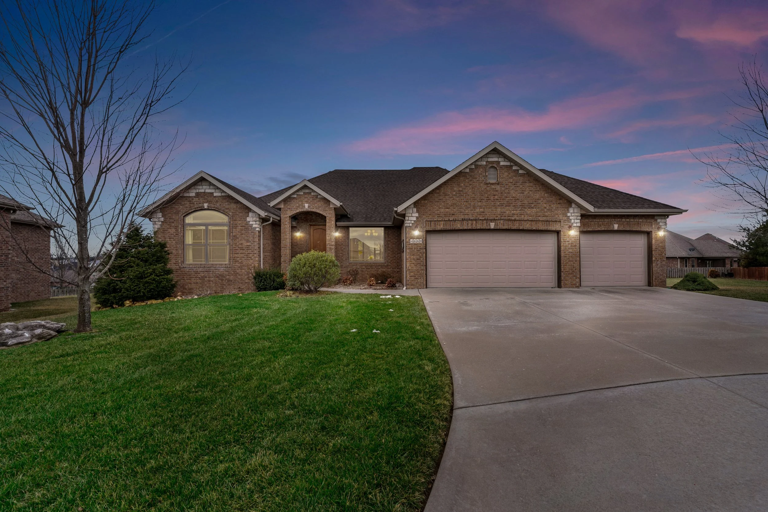 A brick house with attached garage, front yard with green grass, leafless trees, and a sunset sky with pink and purple clouds.