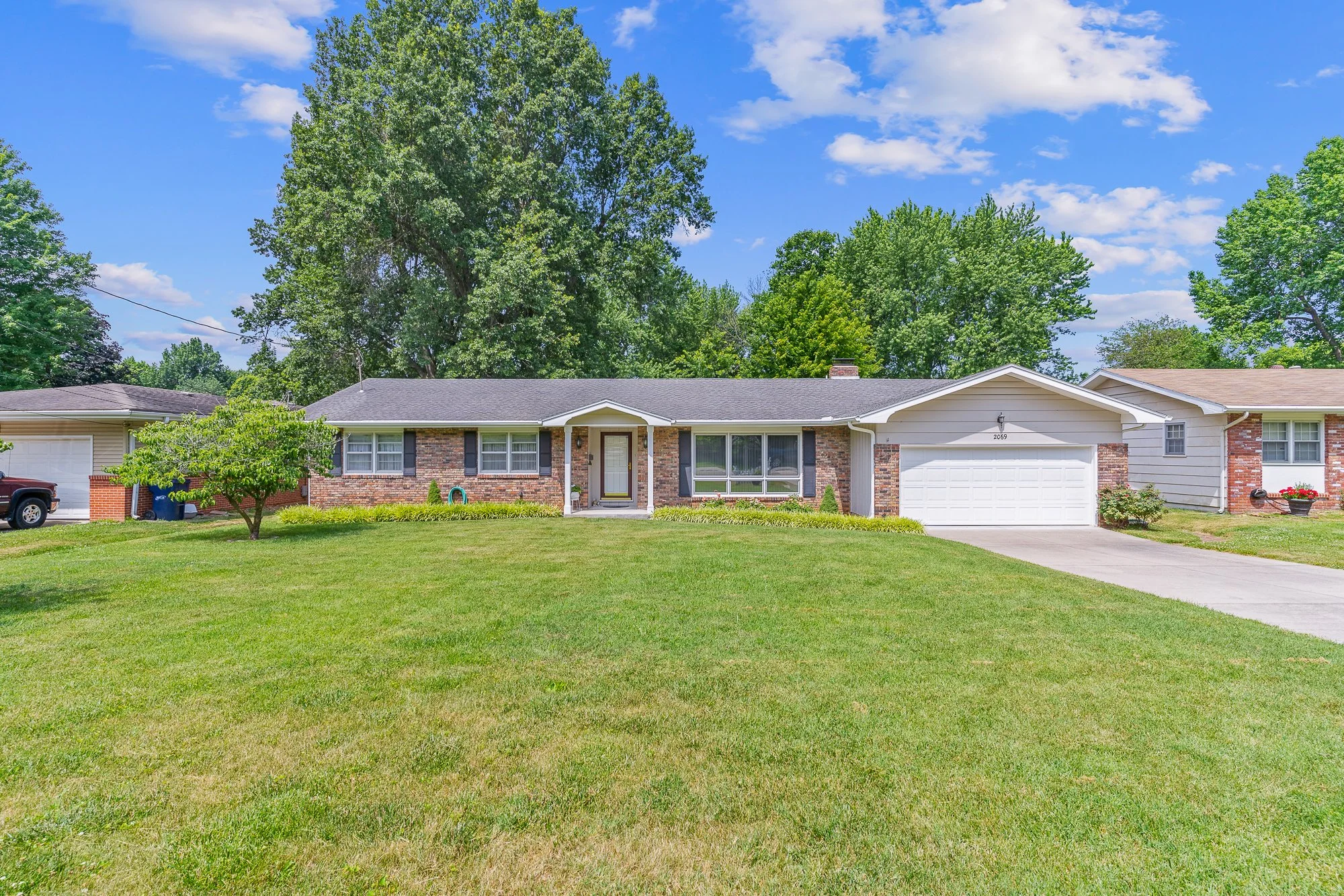 Front view of a suburban house with a manicured lawn, a small tree, and a bright blue sky with some clouds.