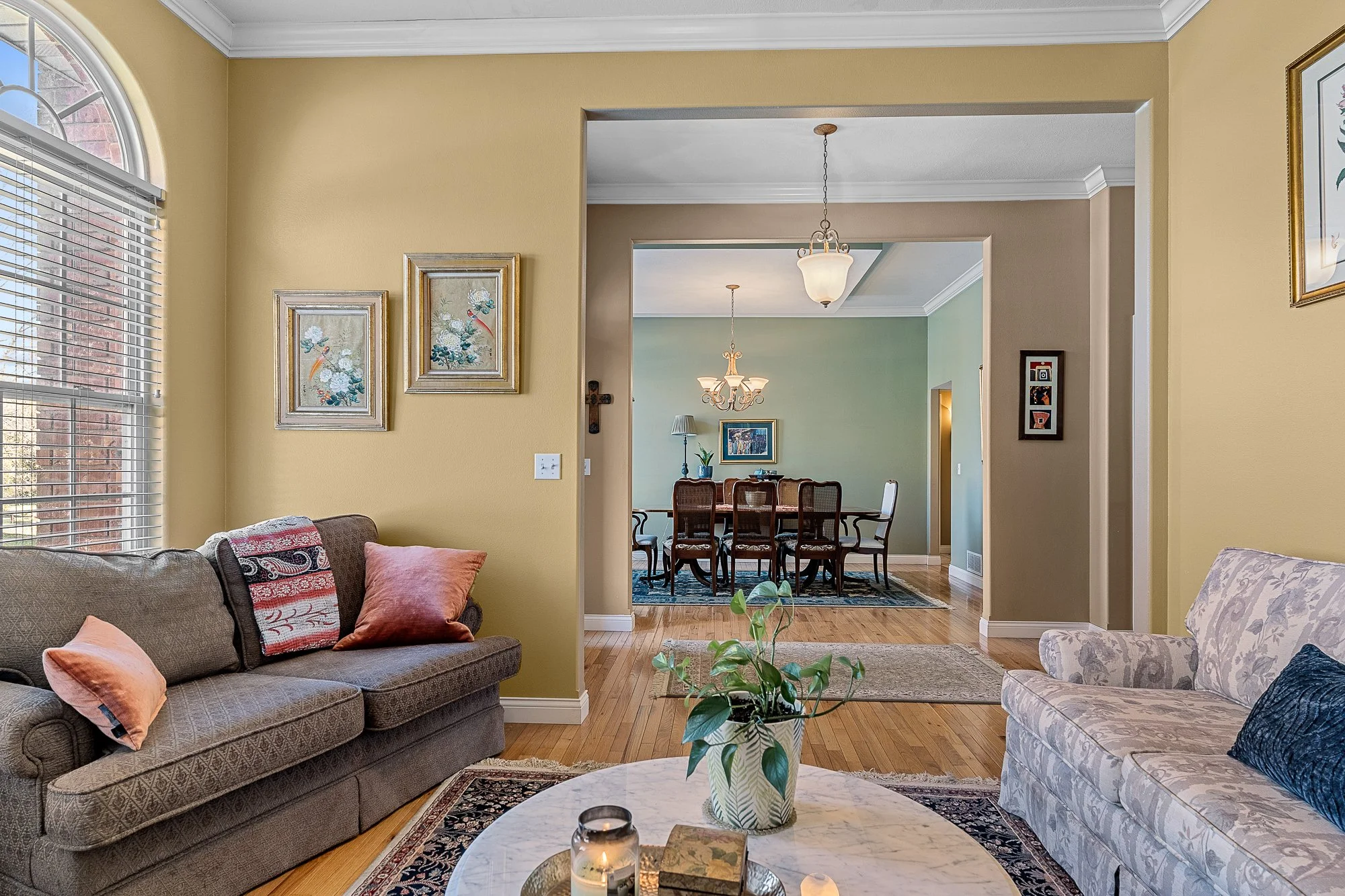 Living room with sofas and decorative pillows, a coffee table with candles and books, a plant, visible in the background are the dining area with a large black dining table and chairs, chandeliers, framed artwork, and light-colored walls.