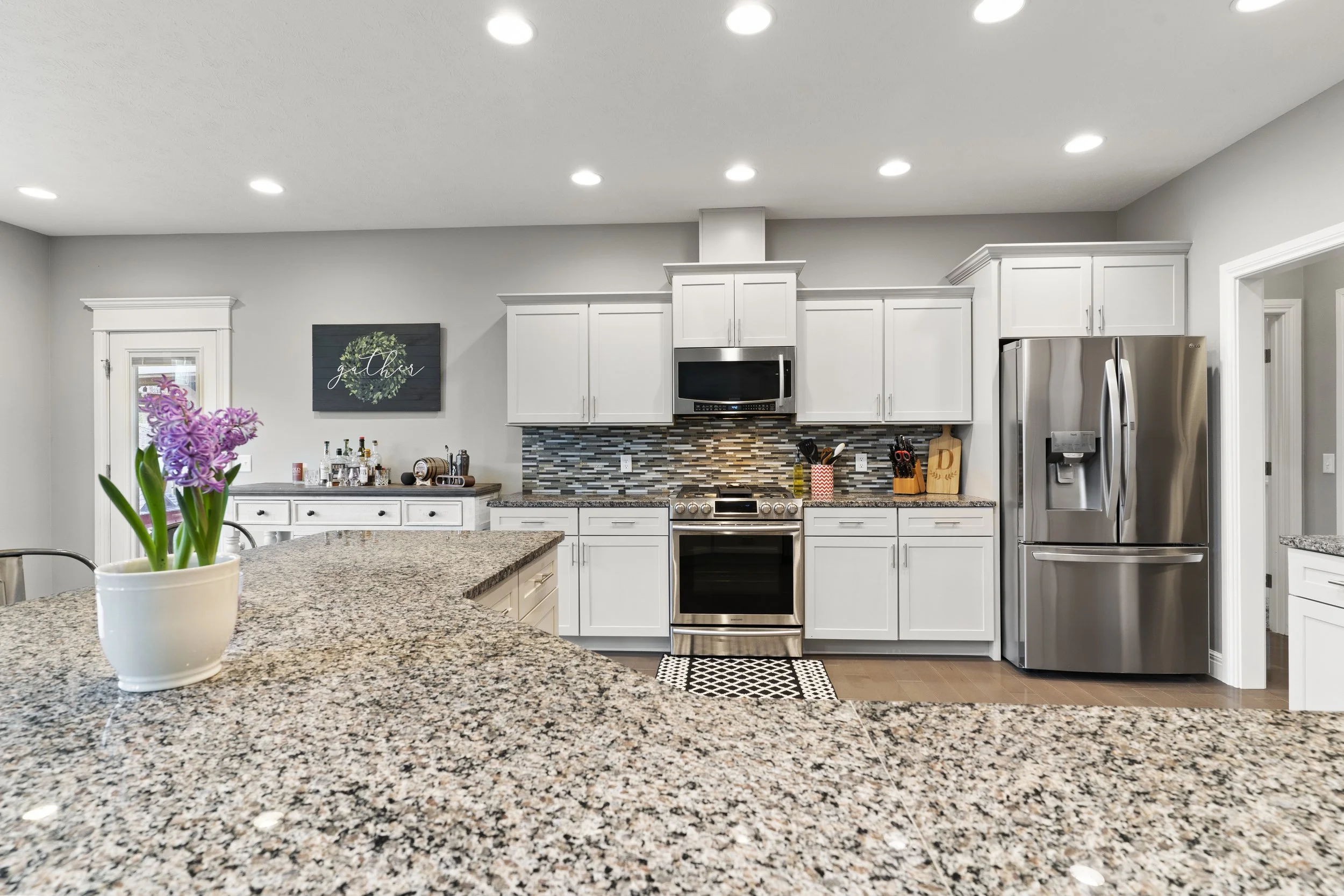 Modern kitchen with white cabinets, stainless steel refrigerator, microwave, and oven, granite countertops, and a mosaic tile backsplash. A purple orchid in a white pot is on the granite island, and a wall decoration reads 'gather' with a wreath desi