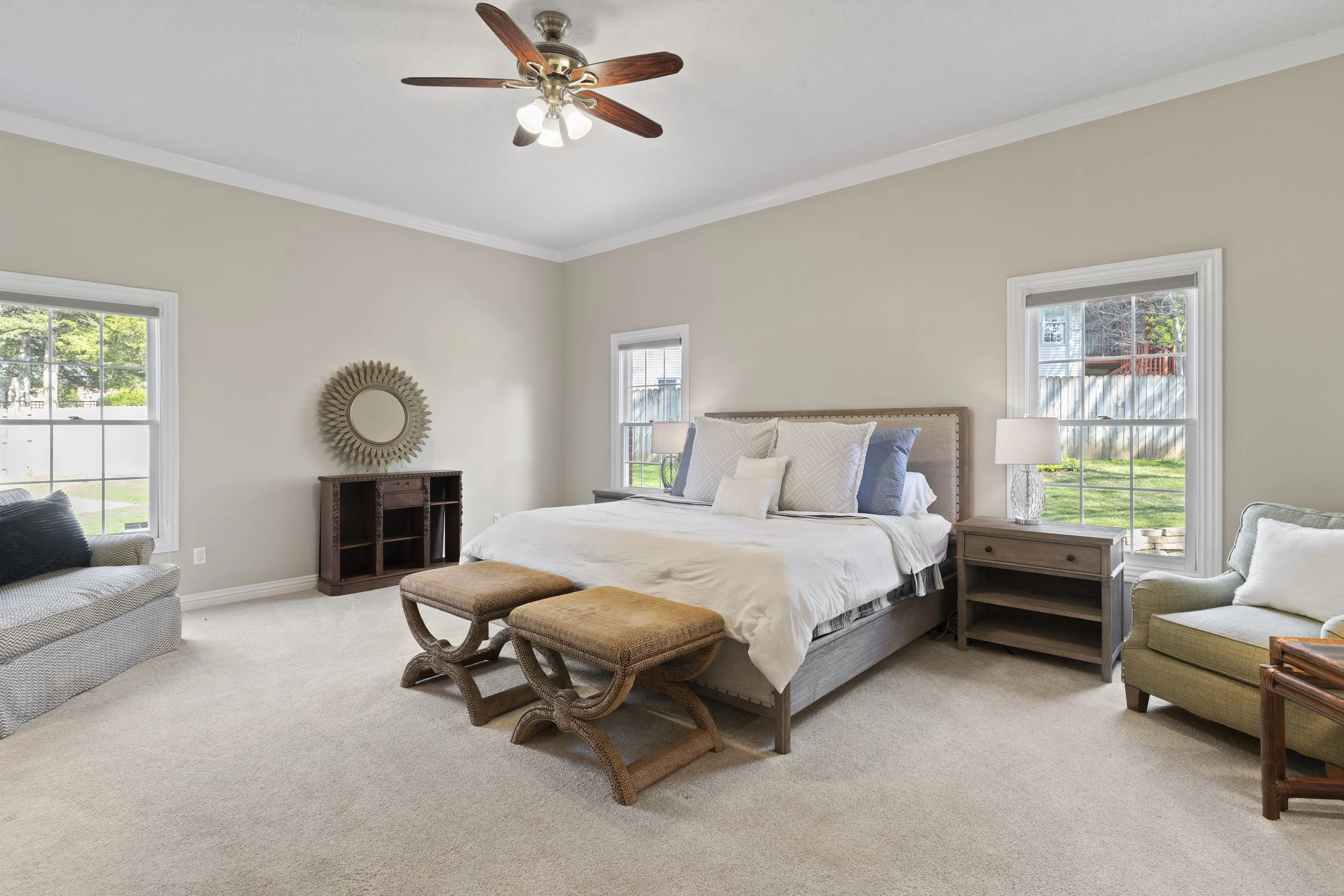 A bedroom with a bed, two windows, two nightstands with lamps, an armchair, and a ceiling fan. The walls are painted a light beige color, and the room is bright with natural light.