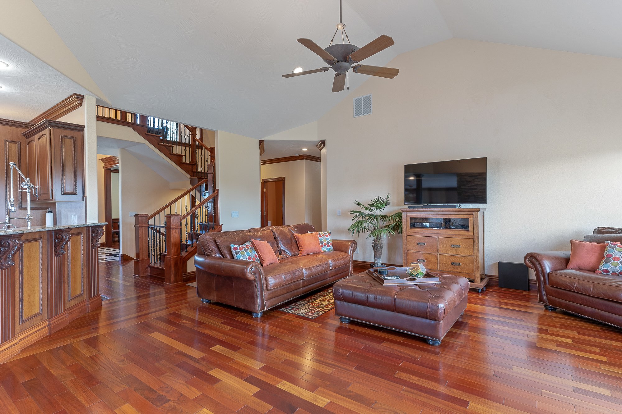 Living room with leather sofas, colorful cushions, a wooden cabinet with a television, a chimney, ceiling fan, hardwood floor, and staircase.