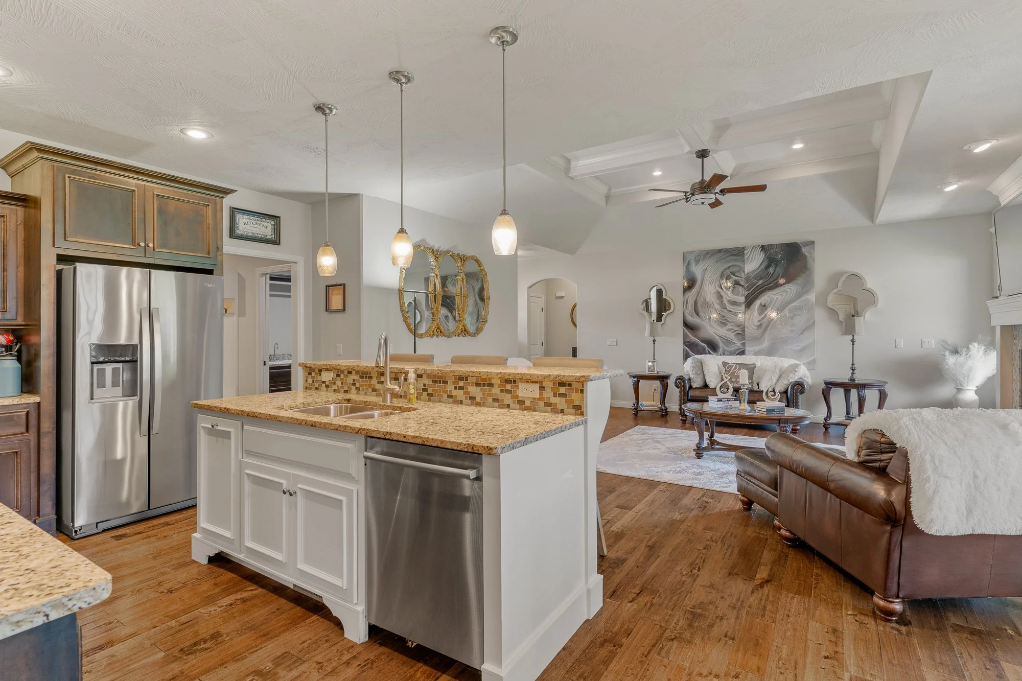 Open-concept kitchen and living room with hardwood floors, white cabinetry, a granite island, stainless steel refrigerator, decorative mirrors, and cozy seating area with a leather sofa and patterned pillows.