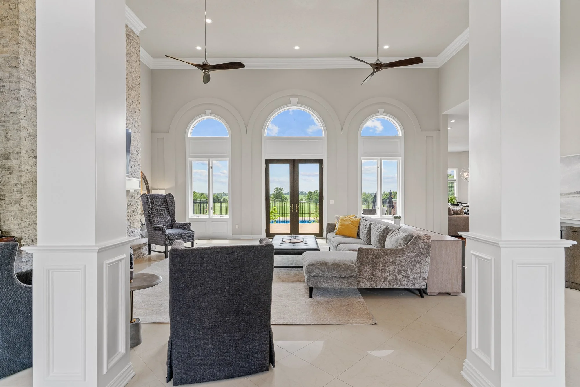 Spacious living room with large arched windows, a sliding glass door, modern furniture including a gray sofa and armchairs, ceiling fans, and a view of a balcony and green landscape outside.