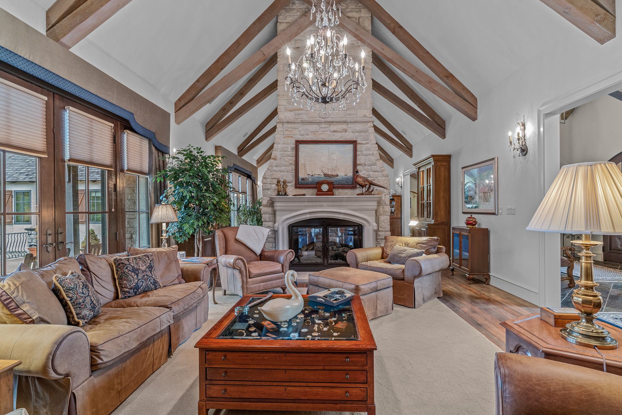 Living room with brown leather sofas, a glass-topped wooden coffee table, a fireplace with a painting above, large windows with blinds, a chandelier, plants, and various decorative items.