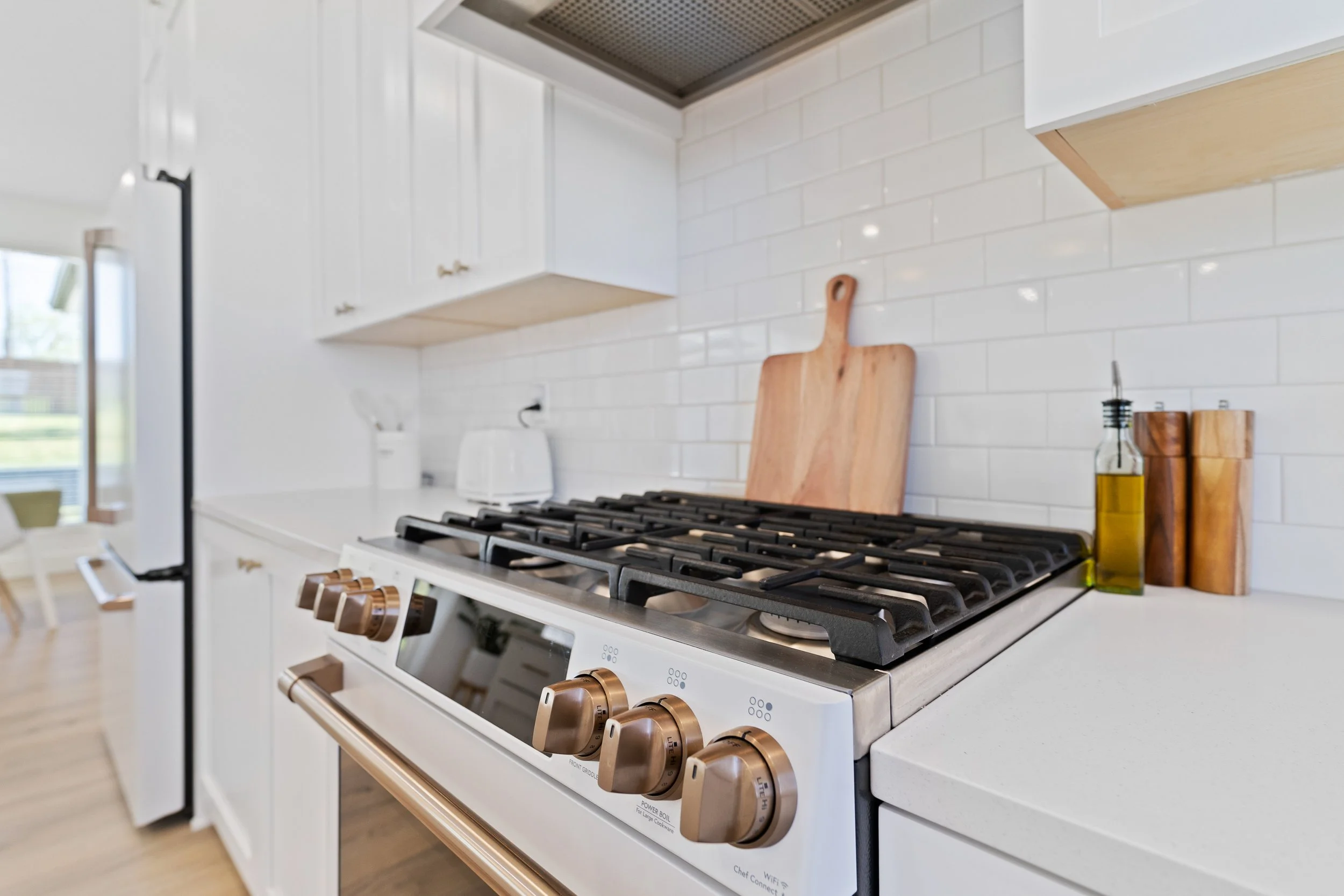 A modern kitchen stove with four burners, white cabinets, a white backsplash, a wooden cutting board leaning against the backsplash, and bottles of olive oil and salt shakers on the counter.