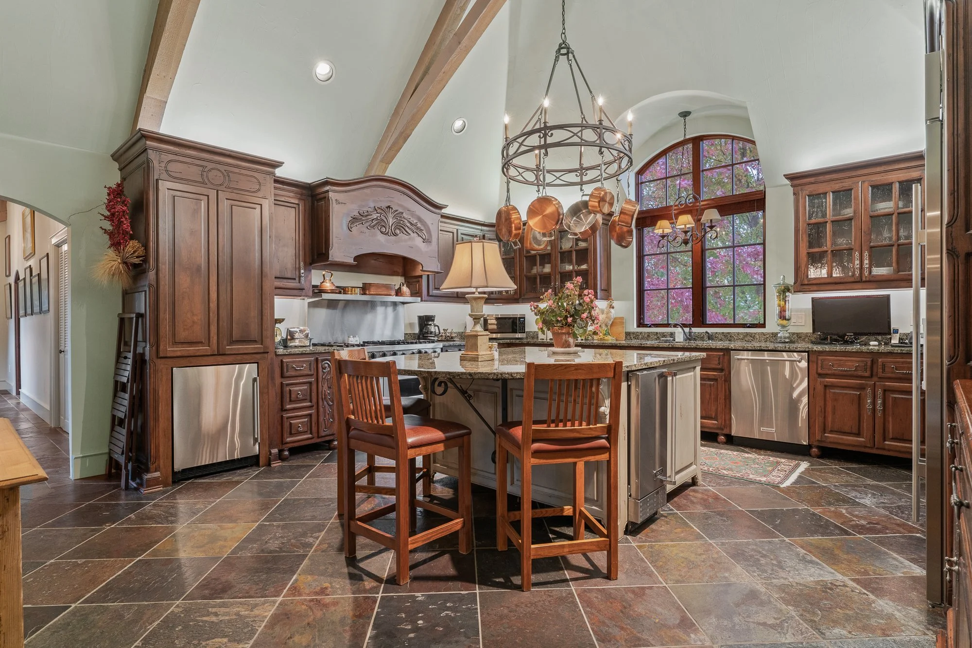 A spacious kitchen with a high vaulted ceiling, wooden cabinets, and a large window overlooking trees with purple and green leaves. A kitchen island with a granite countertop has a flower arrangement and a table lamp. Copper pots hang from a circular