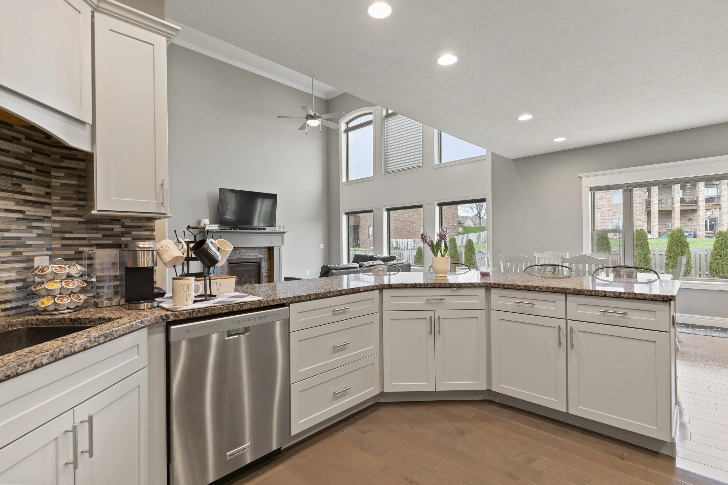 Modern kitchen with white cabinets, granite countertops, a breakfast bar, and stainless steel appliances. Large windows give a view of the backyard.