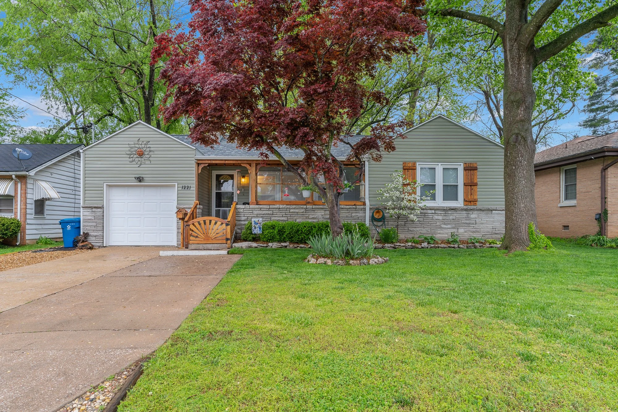 Front view of a house with a driveway, a large red tree, green shrubs, and a lawn. The house has light-green siding, a small porch, and a decorative sun ornament on the wall. Large trees with green leaves surround the house.