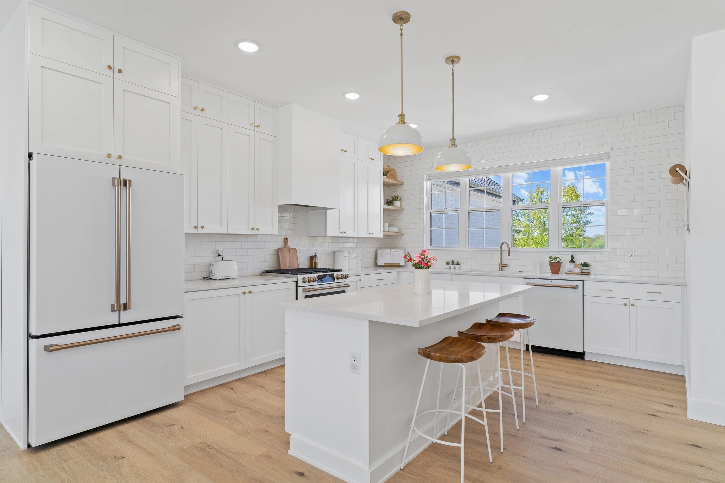 Modern white kitchen with wood flooring, hanging pendant lights, large windows, and a kitchen island with stools.