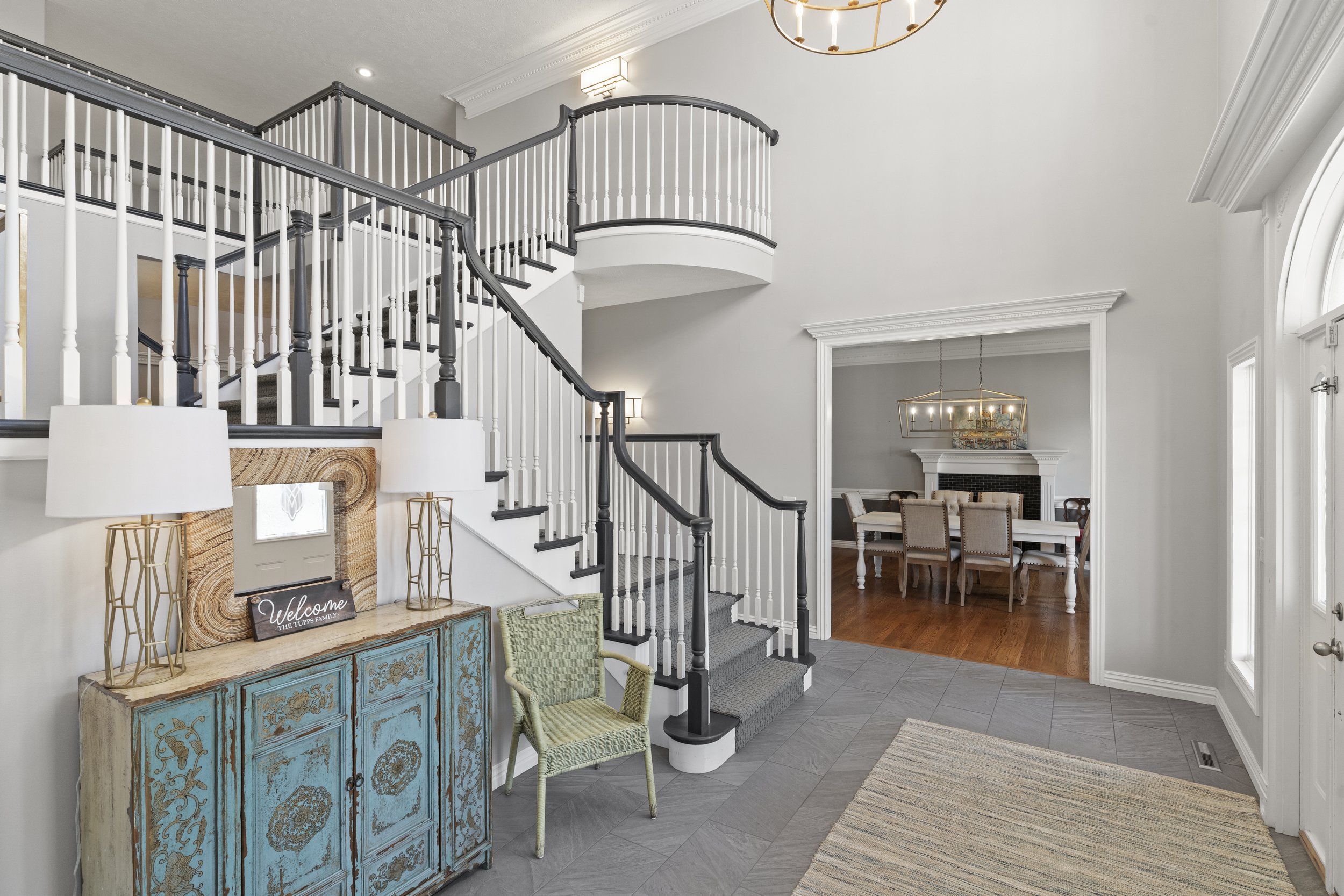 Interior view of a modern home entryway with a staircase, a blue distressed cabinet, two lamps, and a sitting area with a dining table and chairs in the background.