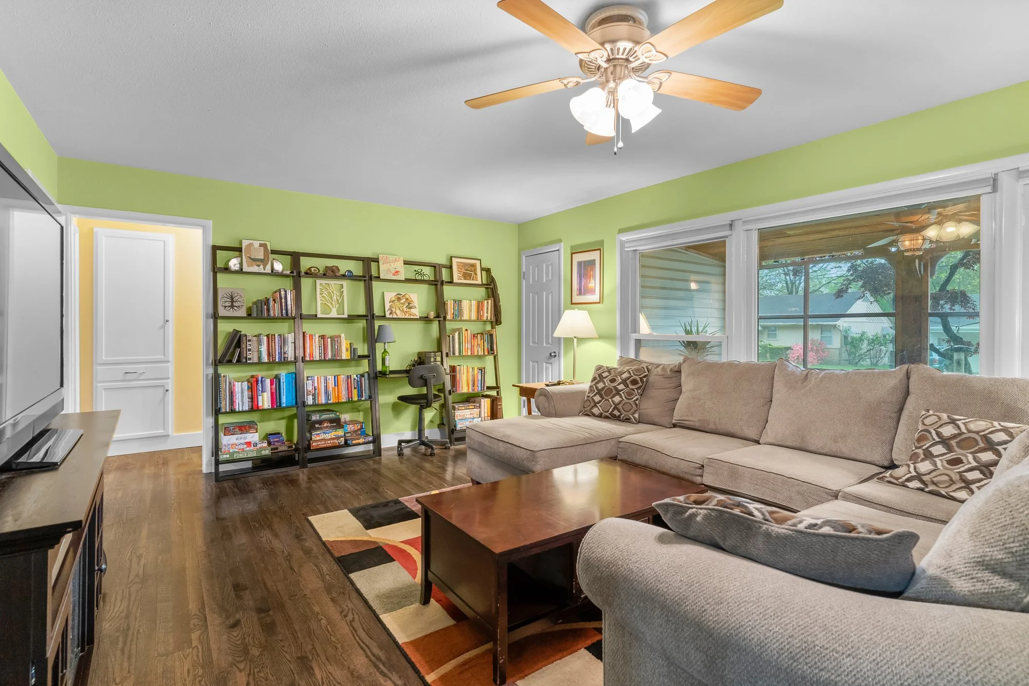 Living room with green walls, beige sectional sofa, wooden coffee table, black bookshelf filled with books, large window showing a porch and trees, ceiling fan with wooden blades and light fixtures, flat-screen TV on a stand, various decorative items