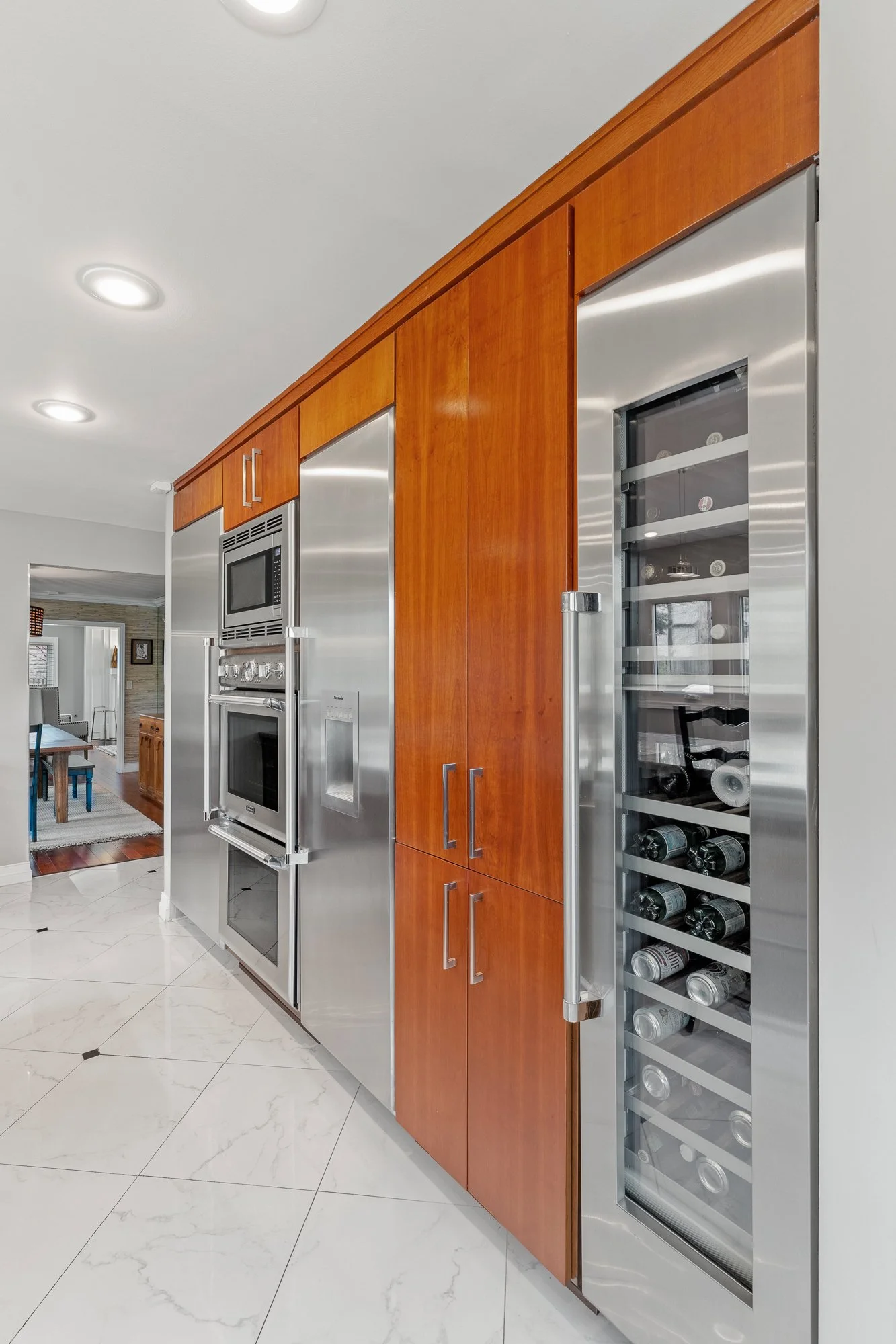 A kitchen with wooden cabinets and built-in stainless steel appliances, including a microwave, oven, and a wine cooler with bottles inside. The floor is tiled in white with black details.