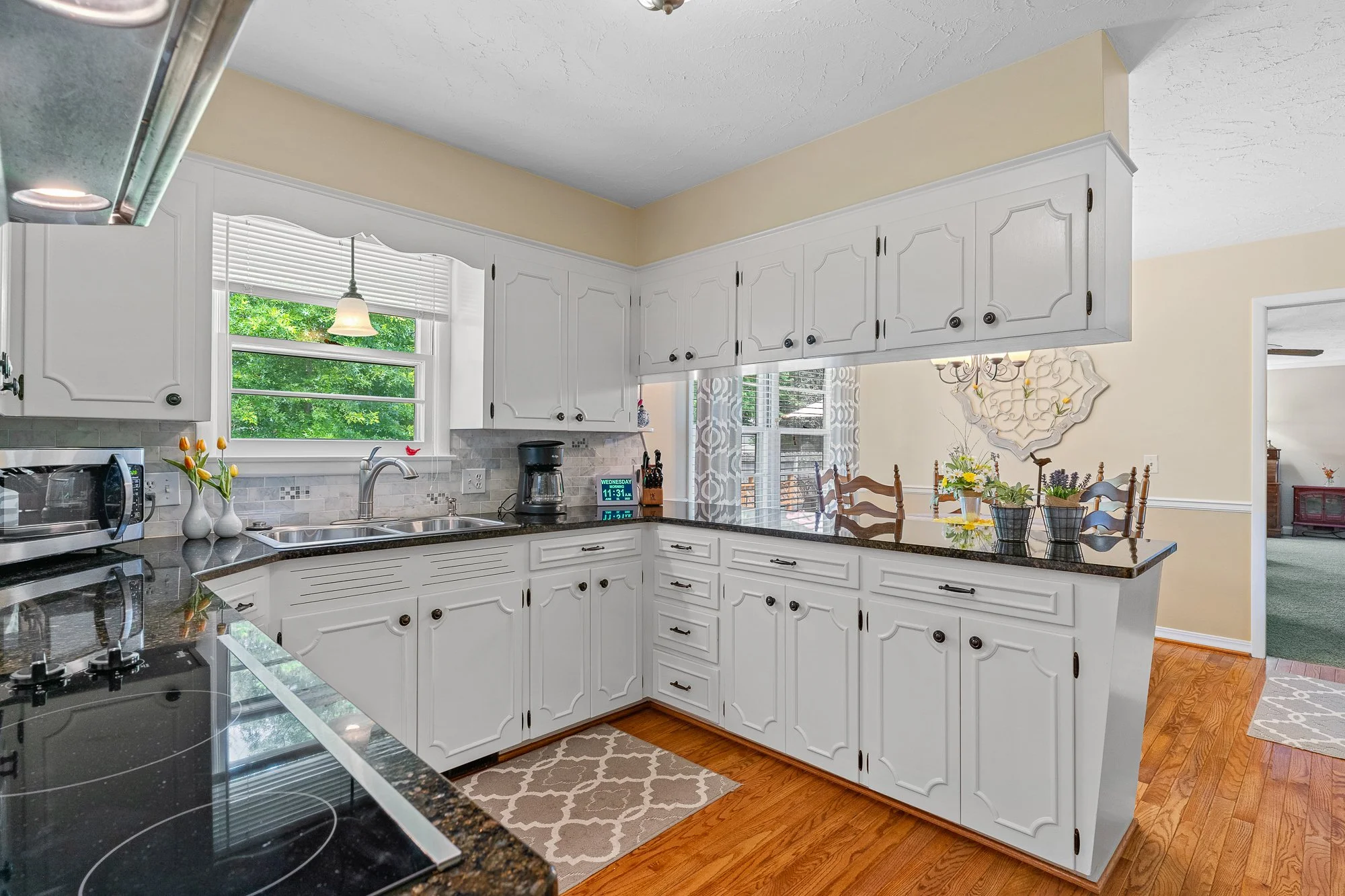 A bright kitchen with white cabinets, a black granite countertop, a window with white blinds overlooking green trees, and a dining area with a wooden table and chairs.