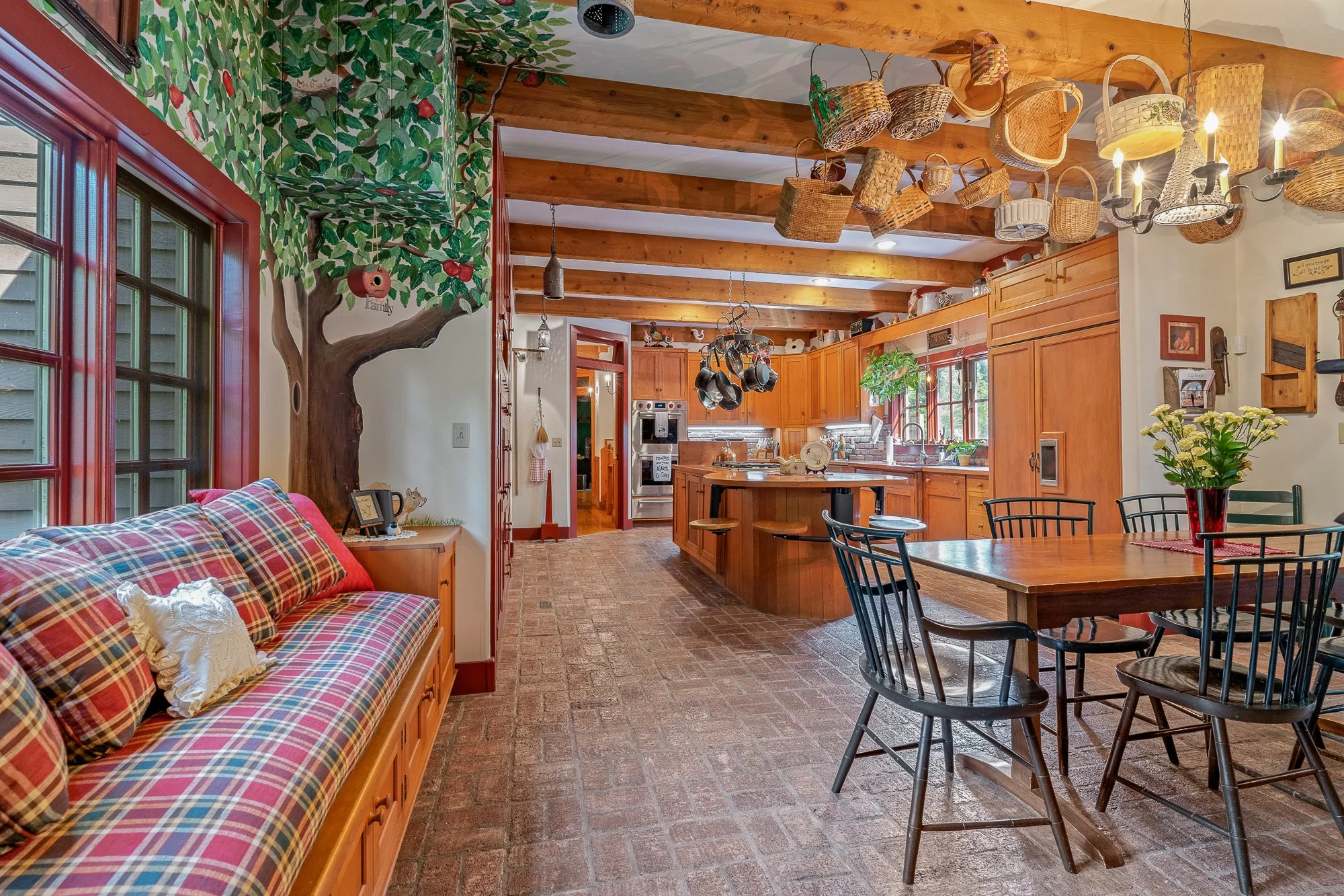 The image shows a cozy kitchen and dining area with a brick floor, wooden cabinets, and a table with black chairs. Decor includes hanging baskets, a chandelier with candles, a painted wall mural of a tree with green leaves and red berries, and a couc