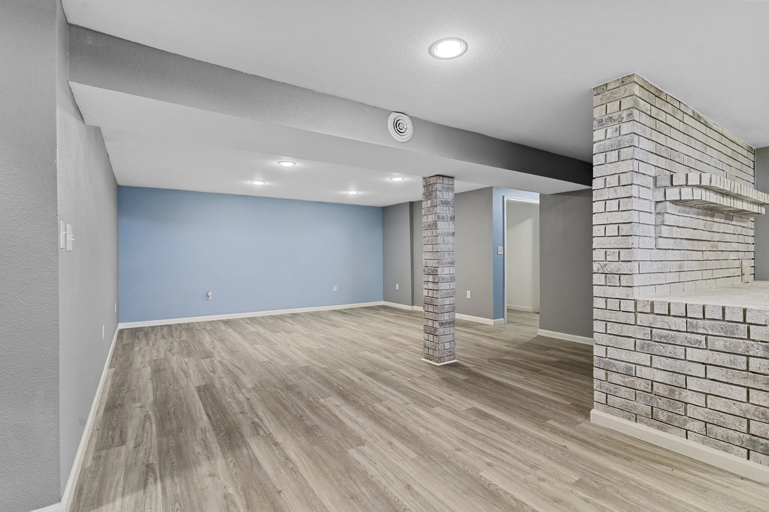 Empty living room with light blue accent wall, gray walls, wood flooring, white baseboards, exposed brick columns, ceiling vents, and recessed lighting.