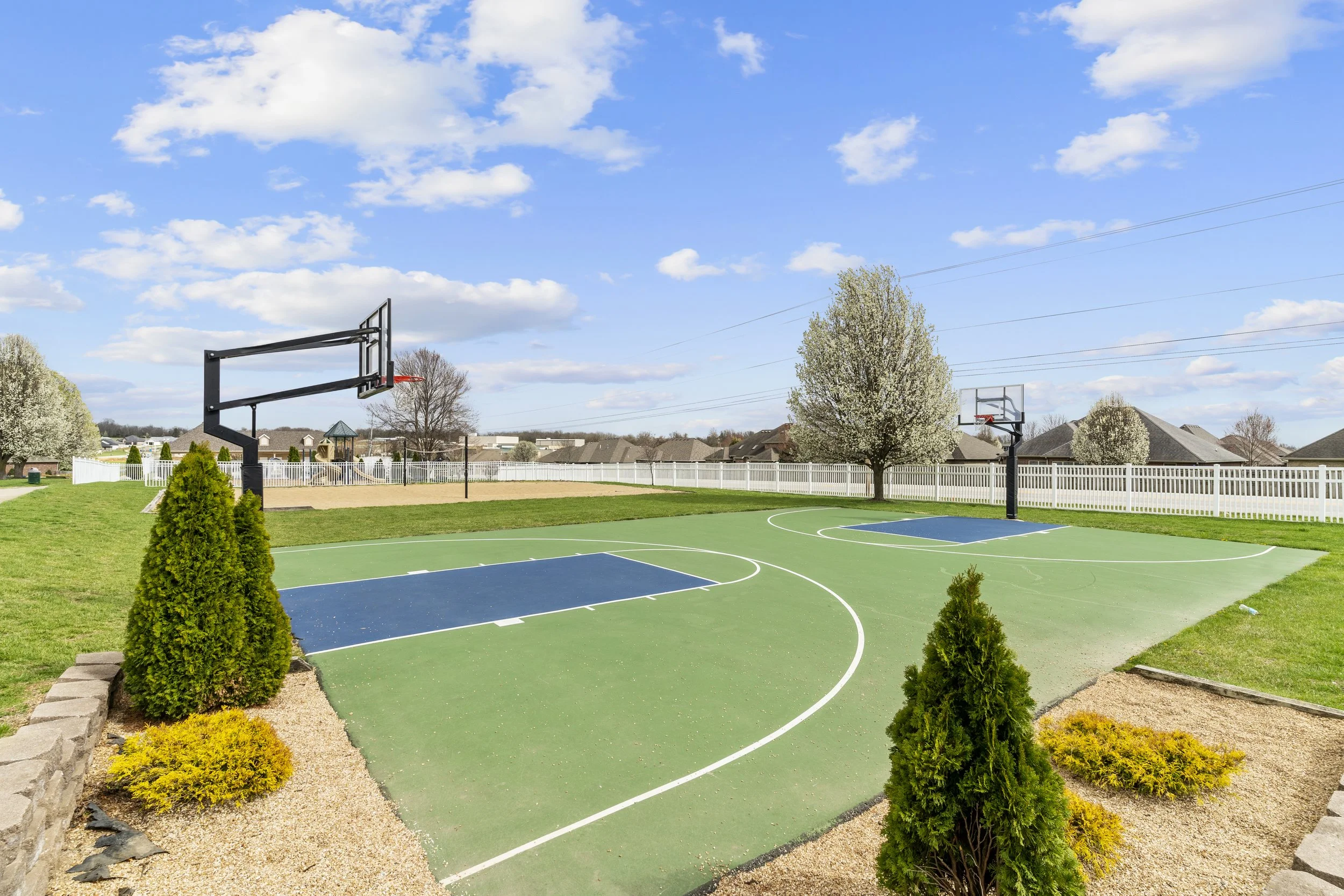 A backyard basketball court with two hoops, surrounded by a white fence and landscaped with bushes and trees, under a partly cloudy blue sky.