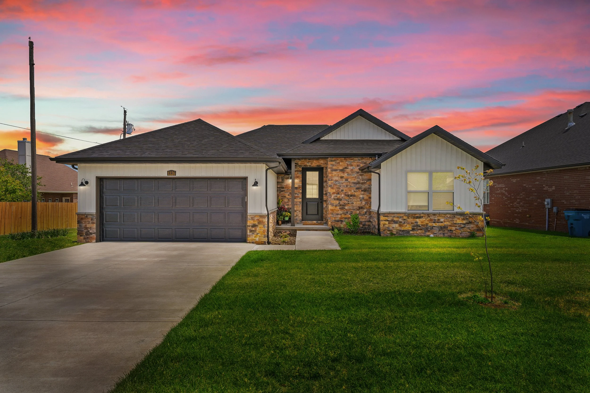 Front view of a modern house with a brick and siding exterior, a two-car garage, a small front lawn, and a colorful sunset sky with pink and orange clouds.