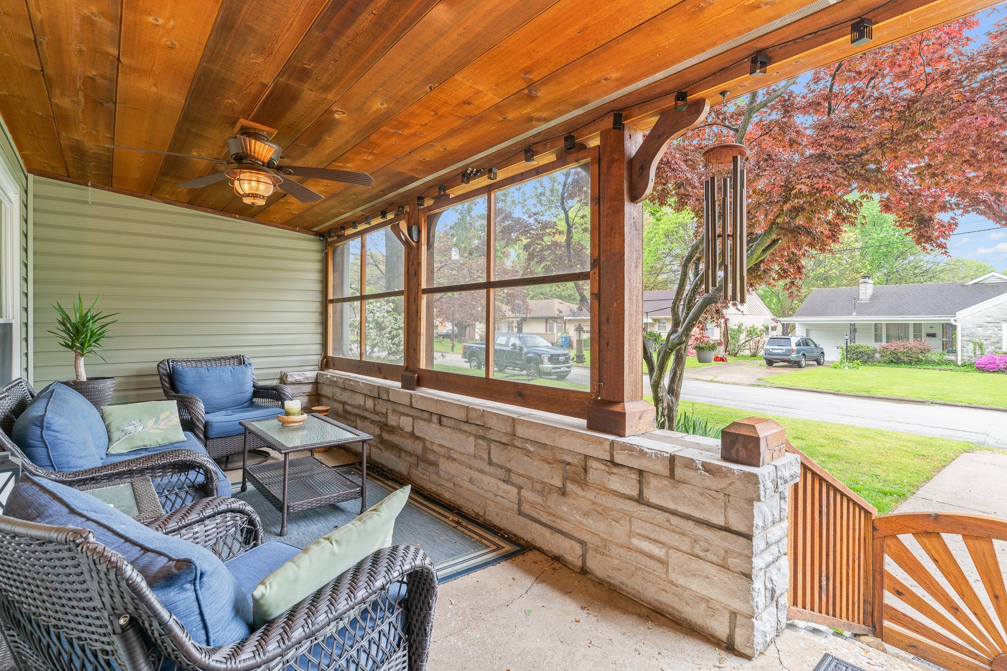 Covered porch with wicker outdoor furniture, blue cushions, potted plant, ceiling fan, wind chimes, and view of a suburban street with trees and houses.