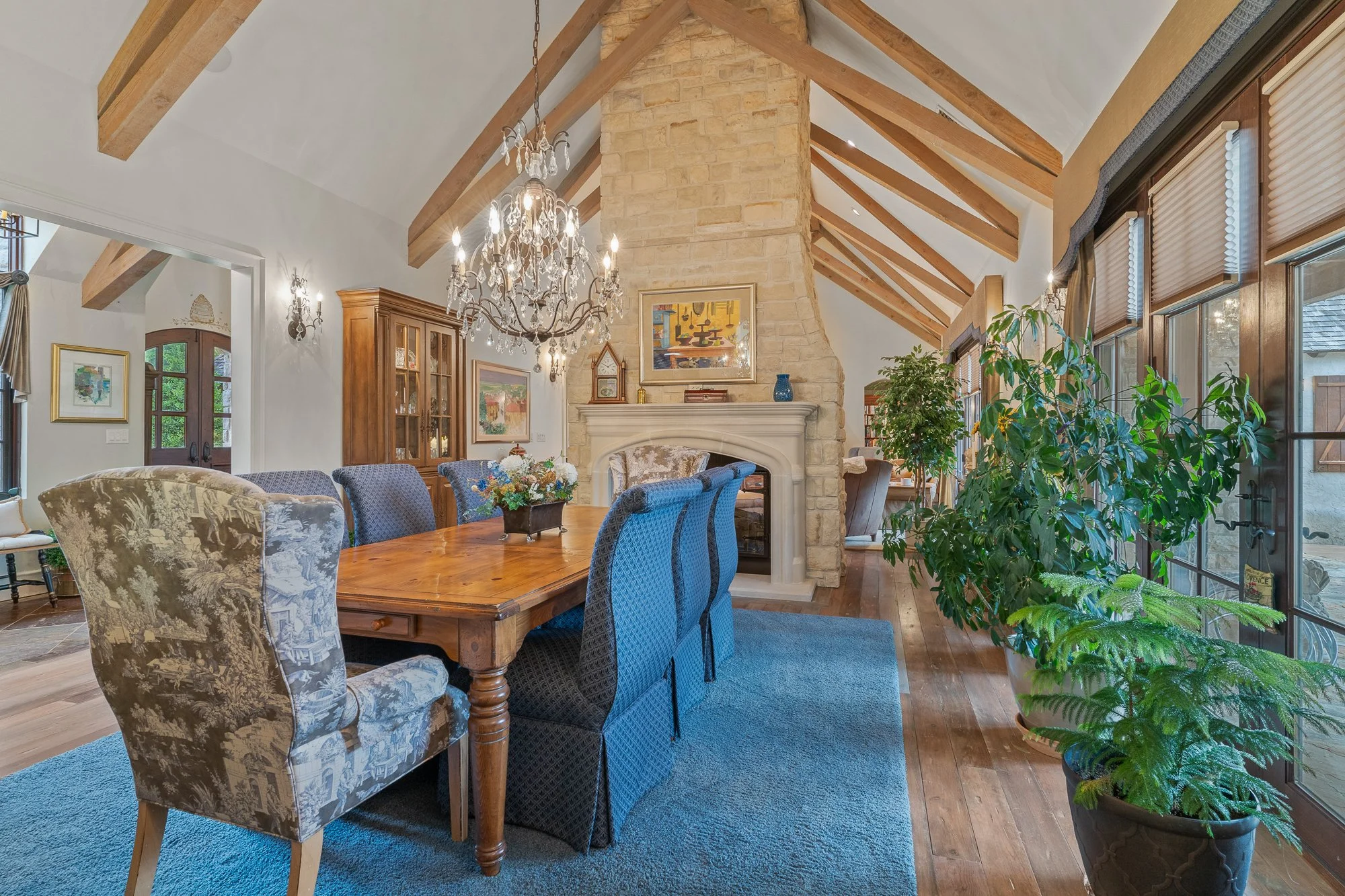A spacious living and dining area with a vaulted ceiling featuring exposed wooden beams. There is a large wooden dining table with upholstered chairs, some with floral patterns and others in solid blue. A chandelier hangs from the ceiling. The room h