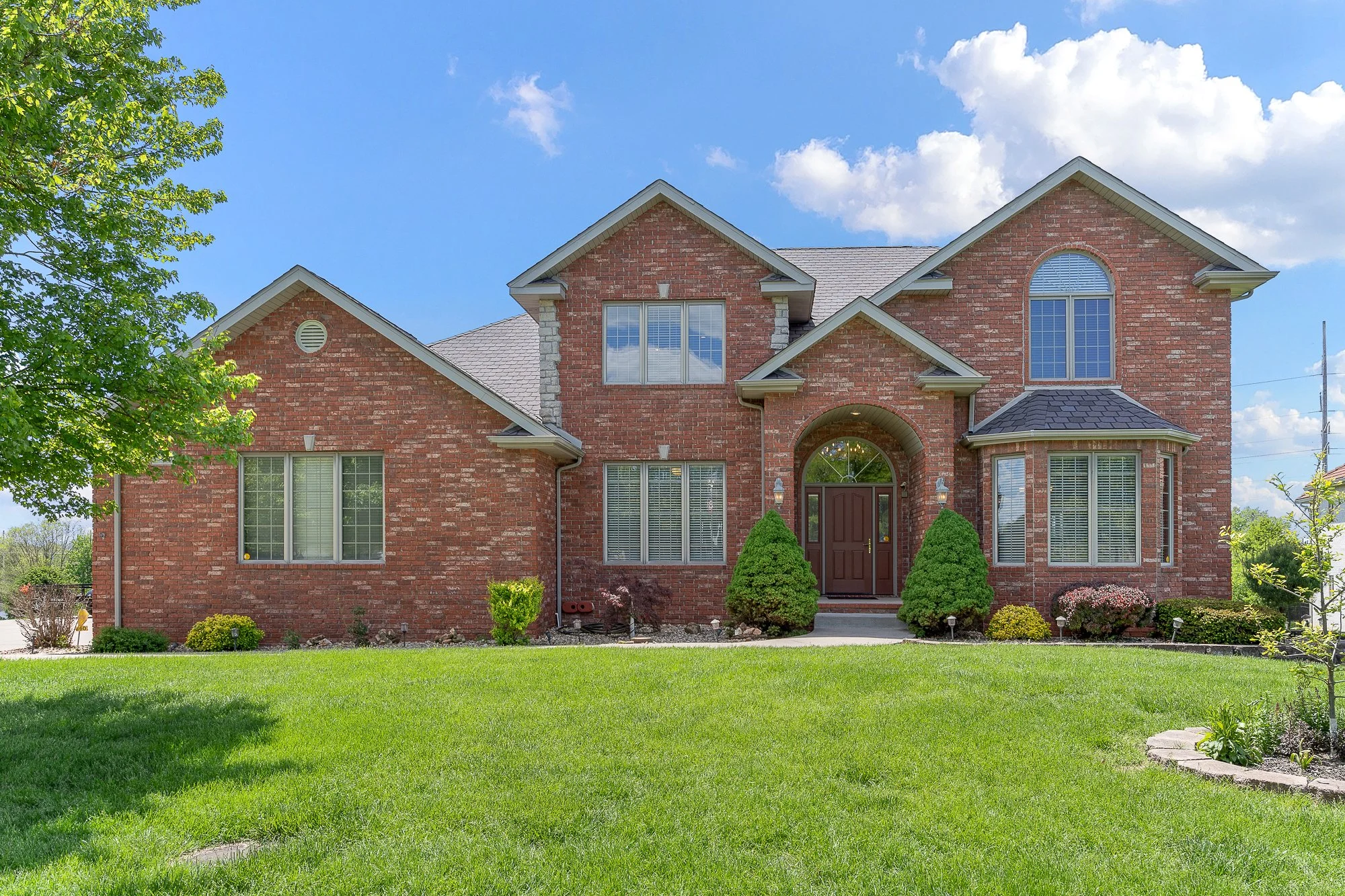A large two-story brick house with multiple gables, white trim, and a front door with a rounded window above it. The house is surrounded by a well-maintained lawn with small bushes and trees, and a blue sky with a few clouds is visible in the backgro